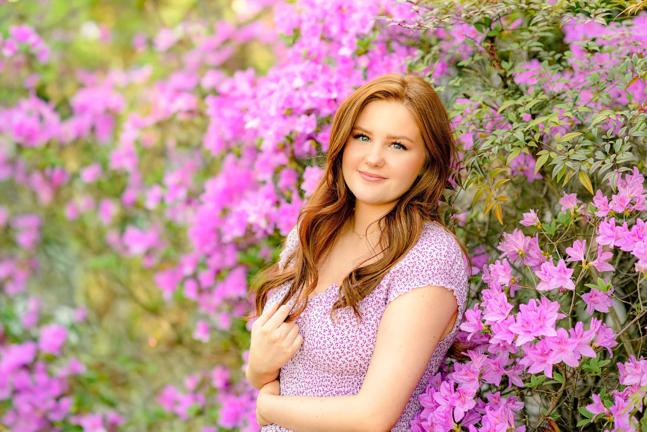 A red-headed senior in a purple flower dress with long, flowing hair, looking directly at the camera with one arm around her waist and the other hand in her hair. She is surrounded by vibrant purple azaleas, showcasing senior photos with flowers.