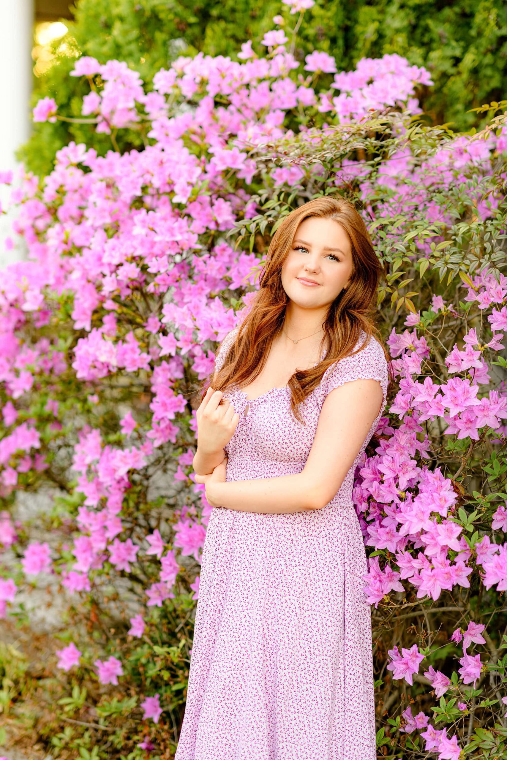 Looking off into the distance with a wistful expression, the red-headed senior stands in front of the blooming azaleas, her hair flowing softly in the breeze, capturing a reflective moment in her senior photos.