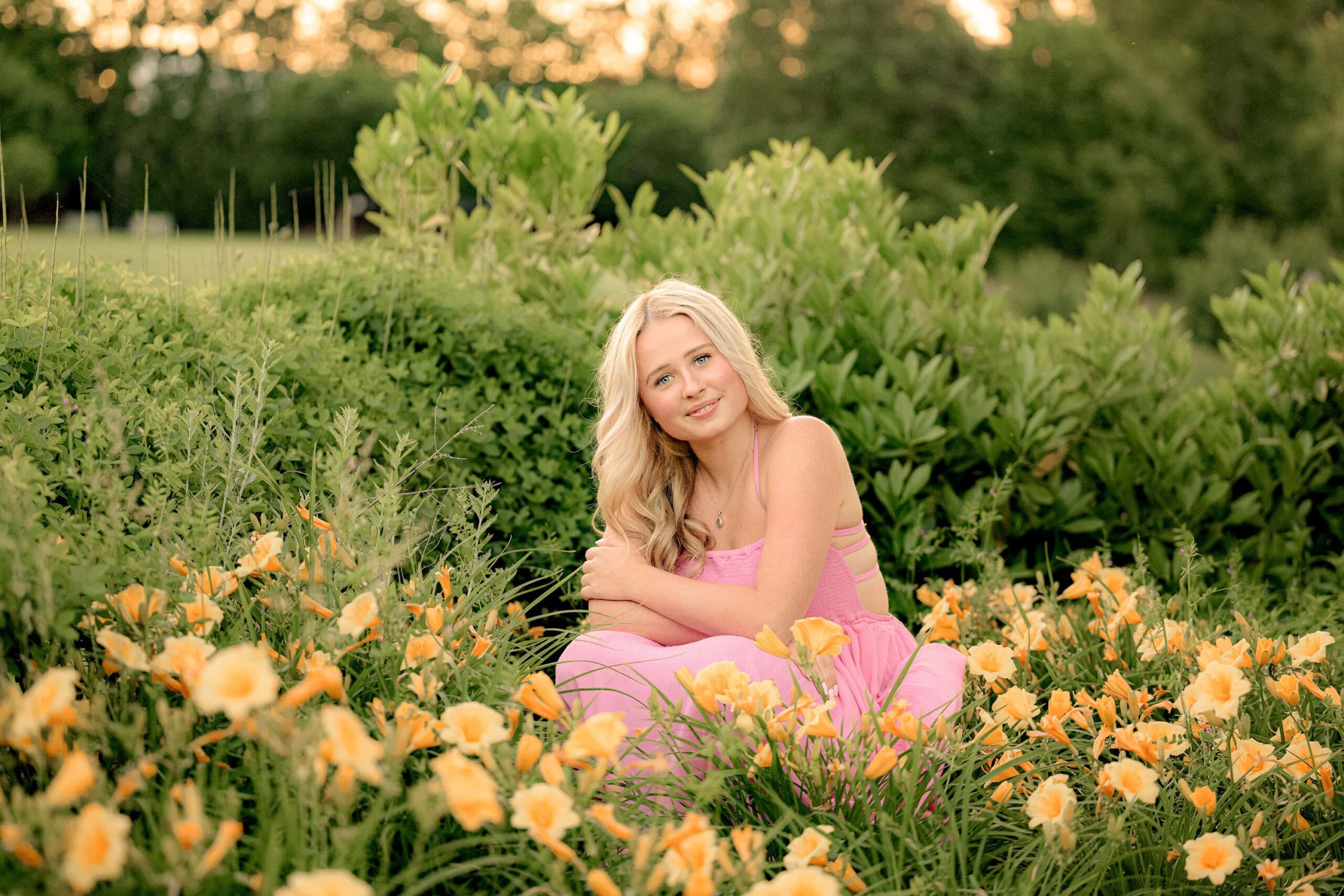 A blonde senior in a pink dress squats gracefully, with golden light shining behind her. She is surrounded by a row of vibrant daffodils in front of her, creating a warm, glowing atmosphere in this beautiful moment.
