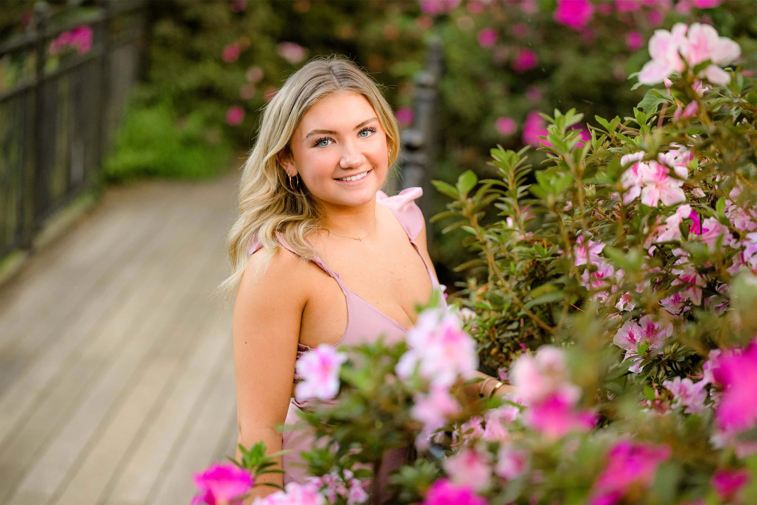 A senior girl poses on a bridge, wearing a pink dress, with a lush bush of pink and purple azaleas in the background. The vibrant flowers complement her dress, creating a beautiful and serene scene.