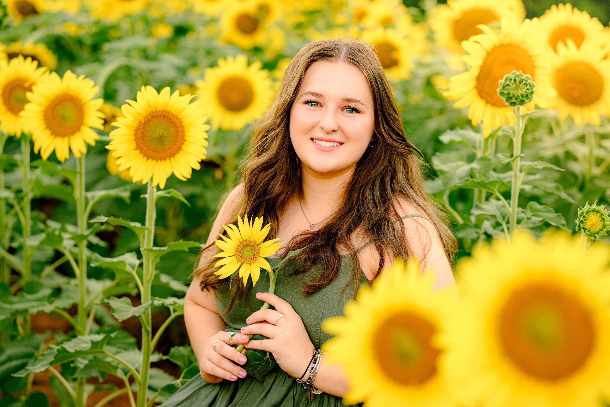 A dark-haired Cuthbertson senior stands in a grove of tall sunflowers, dressed in green, with the golden blooms surrounding her. The vibrant sunflower field is located in the South Carolina area, creating a warm and radiant atmosphere.