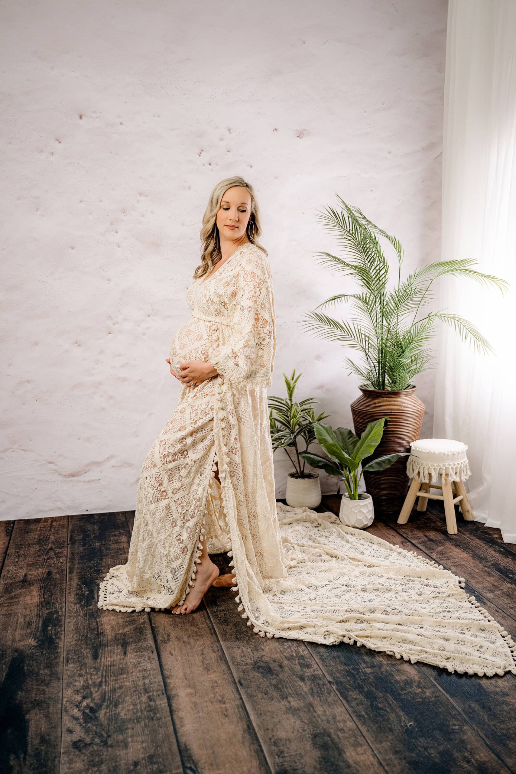 A full-length profile shot of a pregnant mom in a boho-style dress with fringe and side slits, gently cradling her belly with both hands and looking down toward her shoulder. The neutral-toned studio set in Waxhaw features wooden floors and lush green plant accents.
