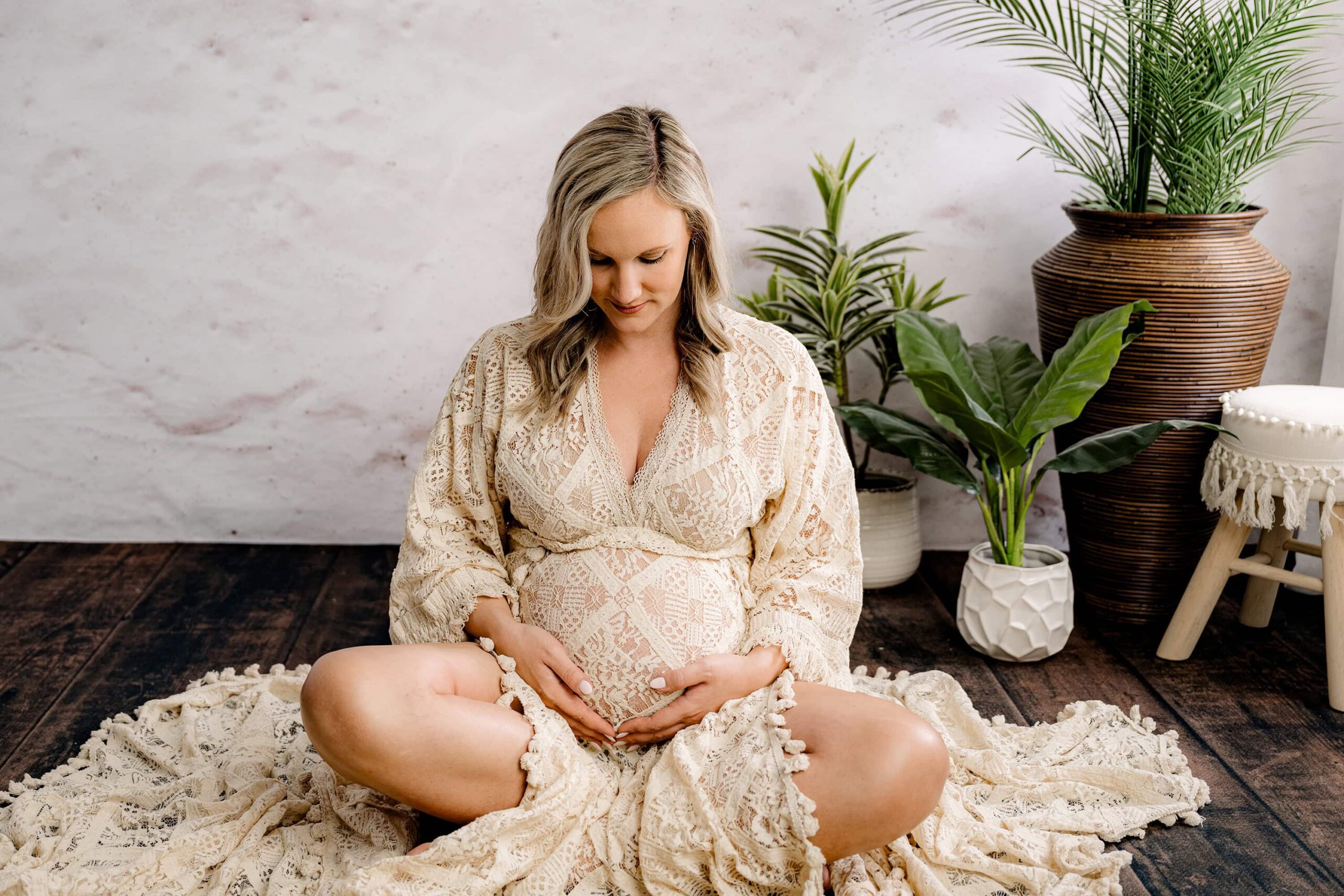 A soft, intimate moment of the expecting mom sitting cross-legged on the wooden floor, looking down at her hands resting on her belly, surrounded by boho-inspired greenery.