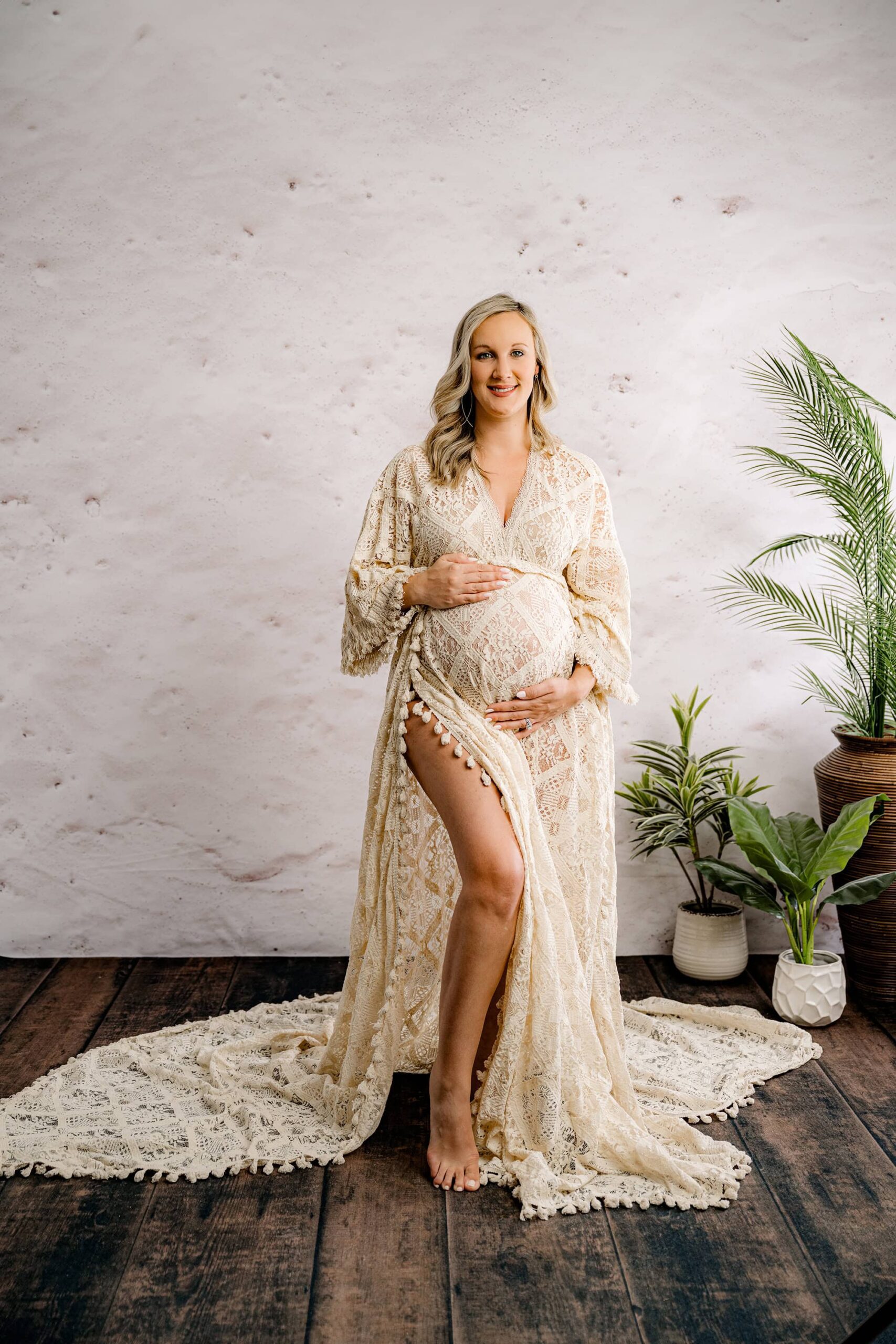 A full-body maternity portrait of the mom standing straight-on, one hand above her belly and one below, with one leg popped slightly. The flowing boho dress complements the earthy studio aesthetic
