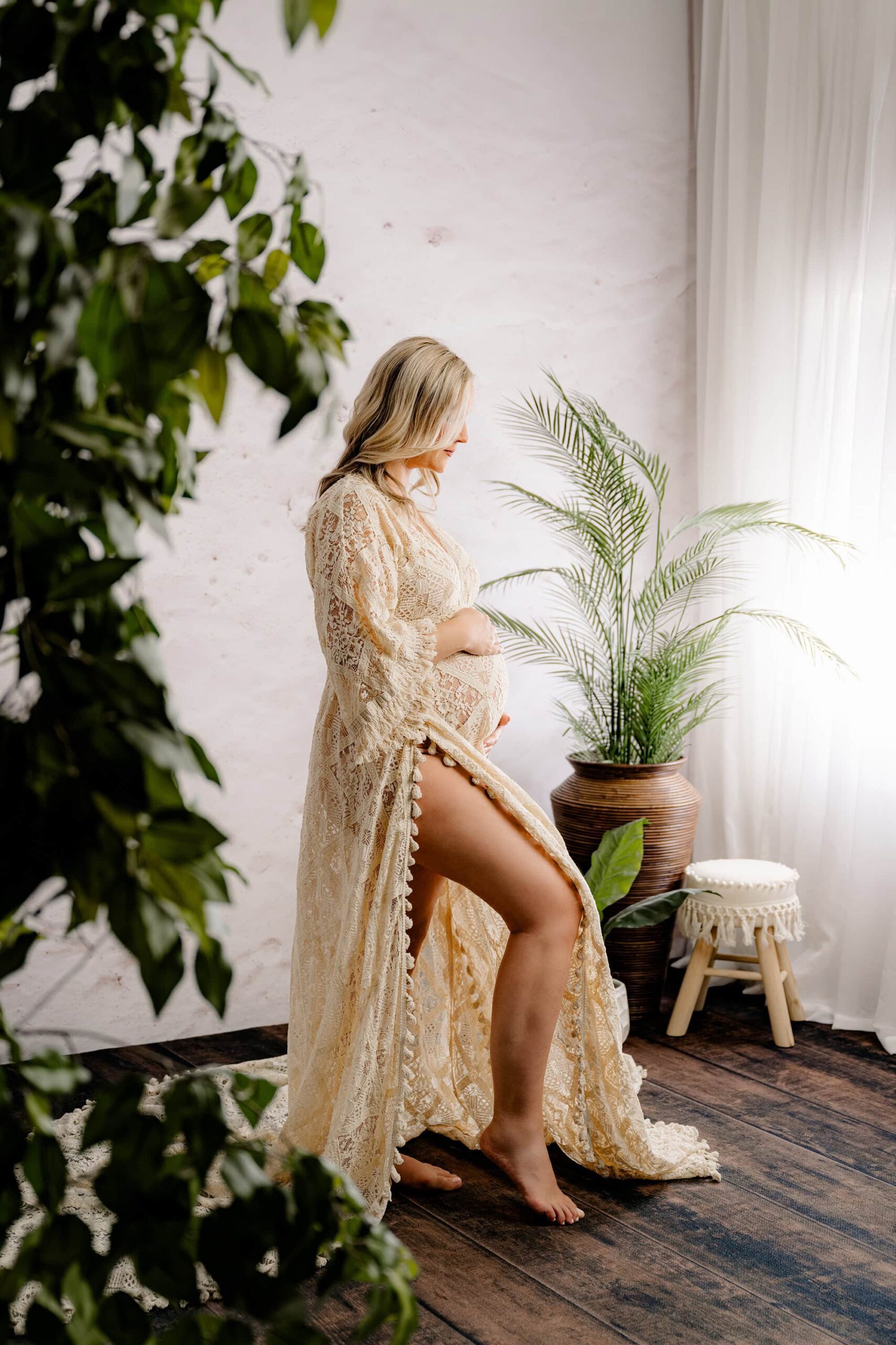 A side-profile shot with a softly blurred plant in the foreground, drawing focus to the mom as she looks down at her belly. A window and boho-inspired decorations in the background add depth and warmth to the composition.