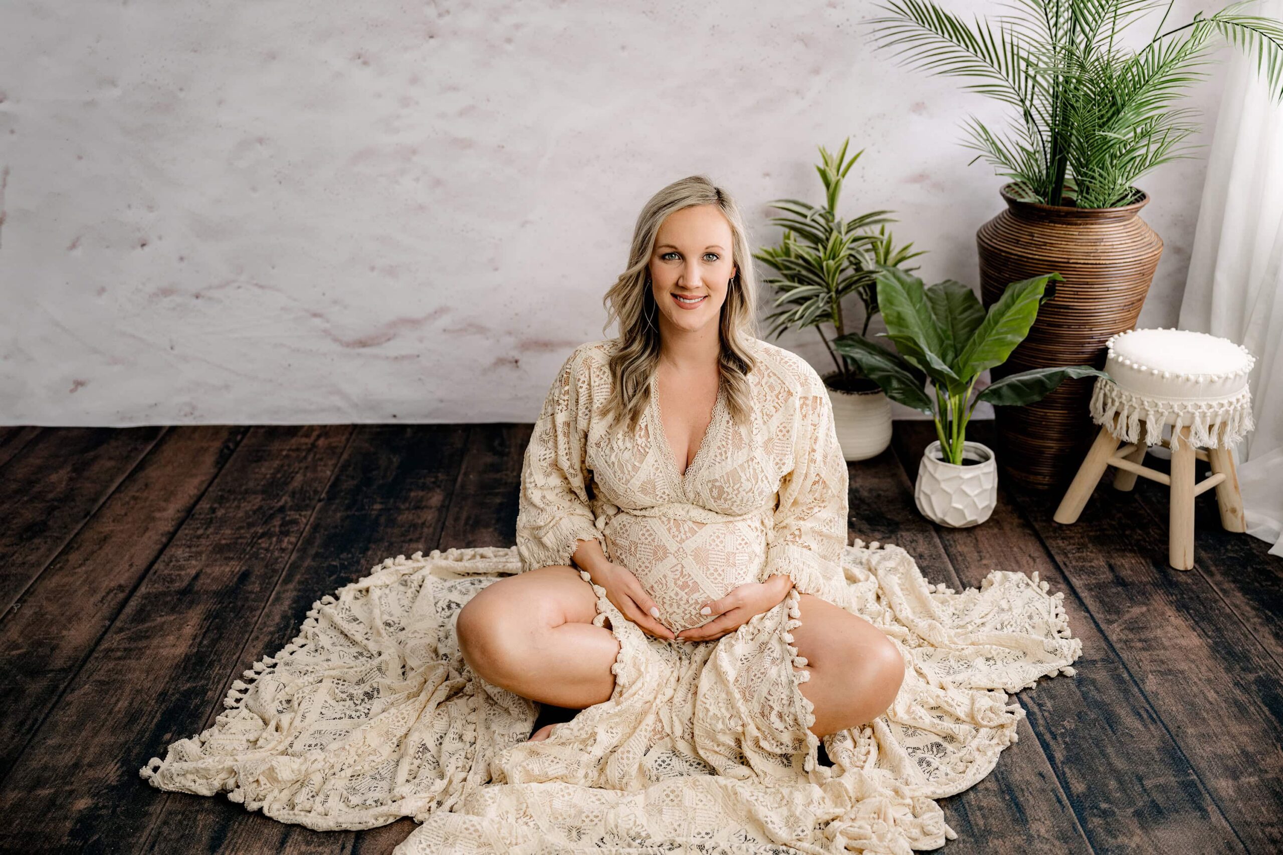 A seated maternity portrait featuring the mom in a client closet boho dress, cross-legged, with both hands gently placed on her belly as she looks directly into the camera with a warm expression.