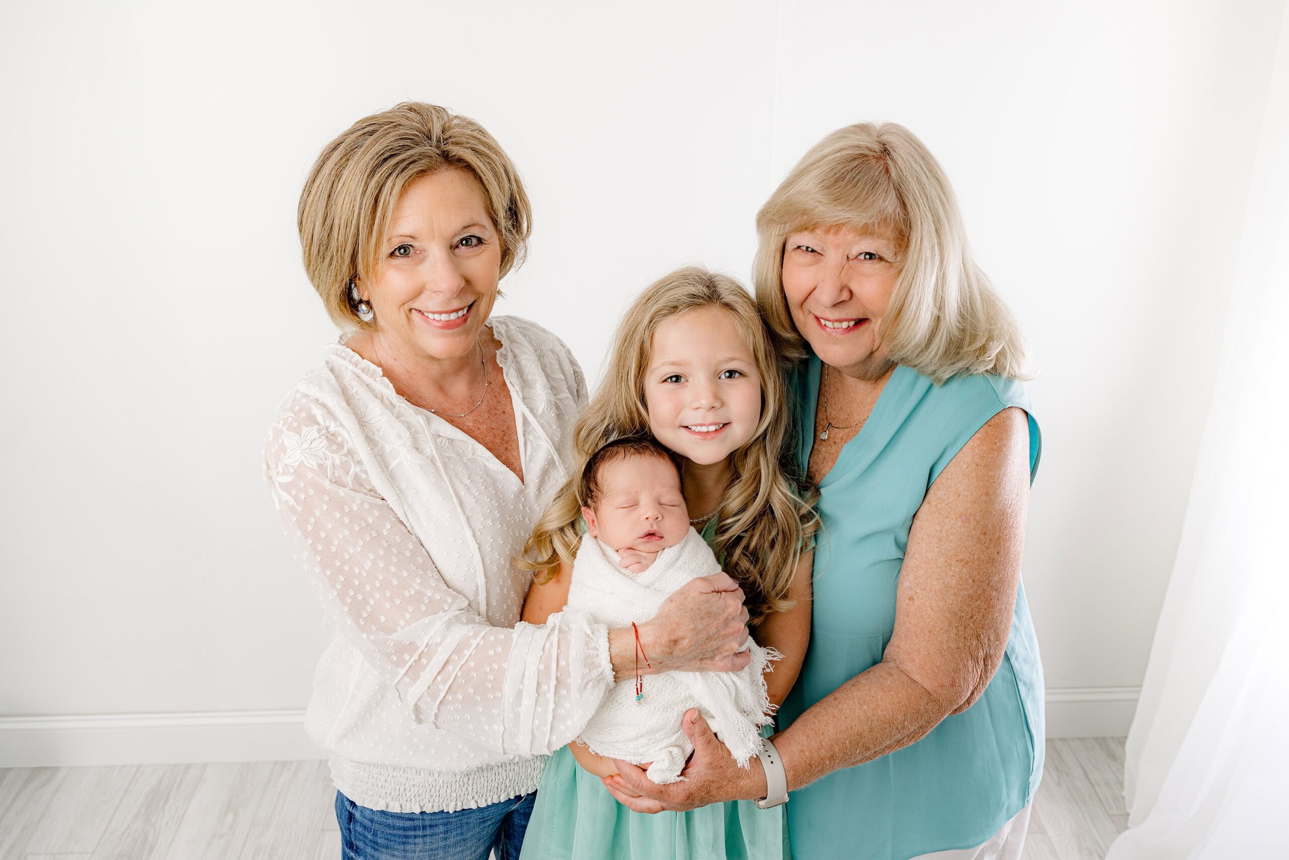 A joyful little girl smiles as she proudly holds her newborn baby brother, wrapped in white, with both grandmothers lovingly supporting her in my Waxhaw, NC studio. This beautiful moment captures the warmth of grandparent love across generations.