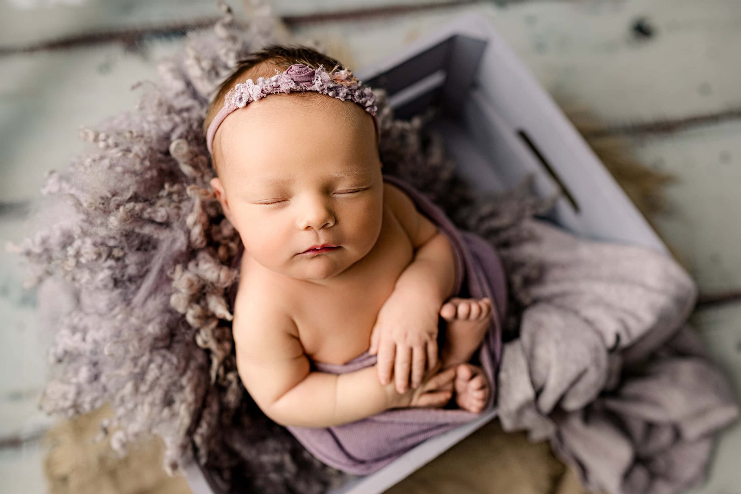 Newborn baby girl, isla, posed in a purple crate surrounded by a purple toned fur and wrapped in a knit wrap in my waxhaw, nc studio.