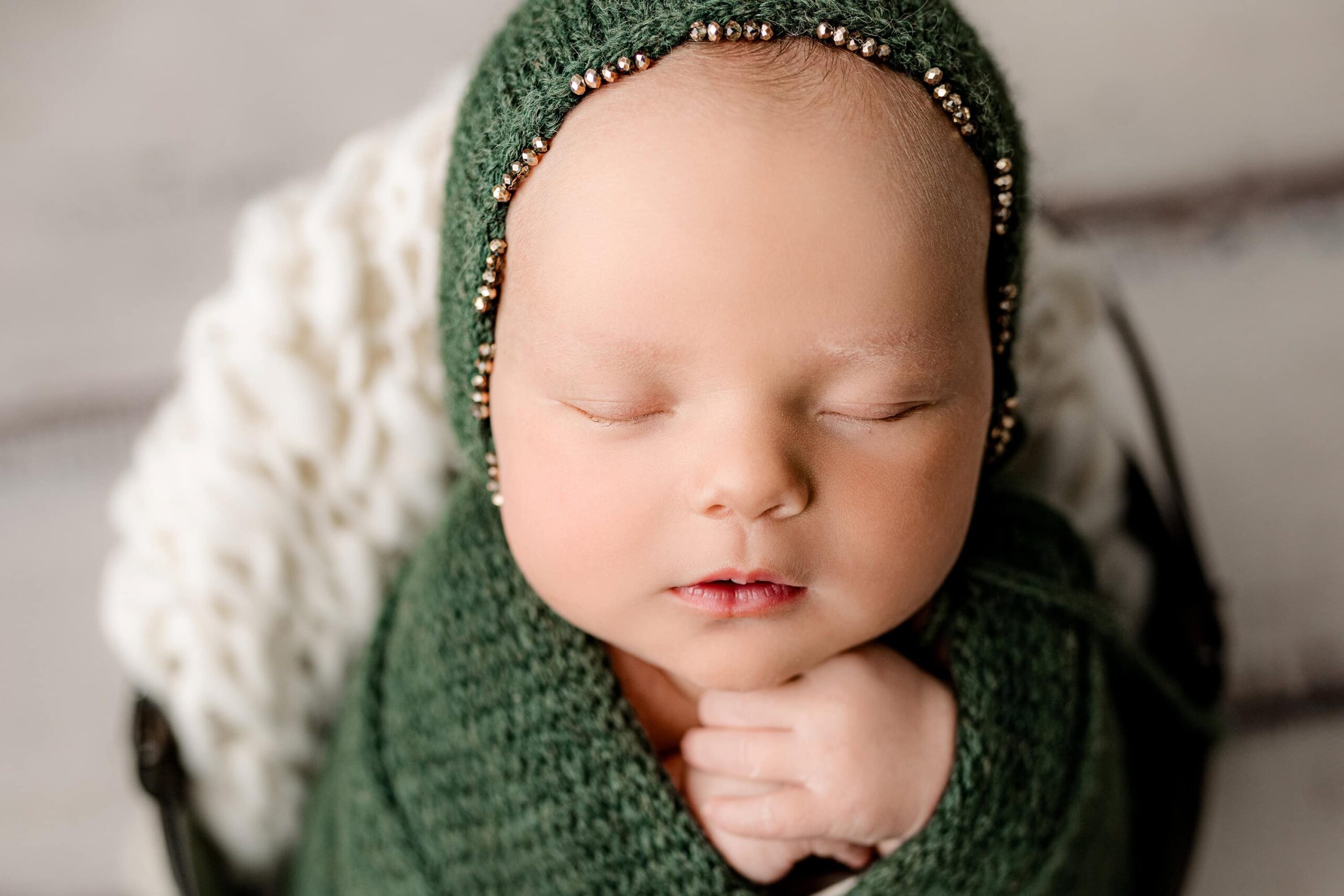 close up image of a newborn baby girl posed with her hand together wrapped in a deep green knit wrap and wearing a green bonnet with gold trim