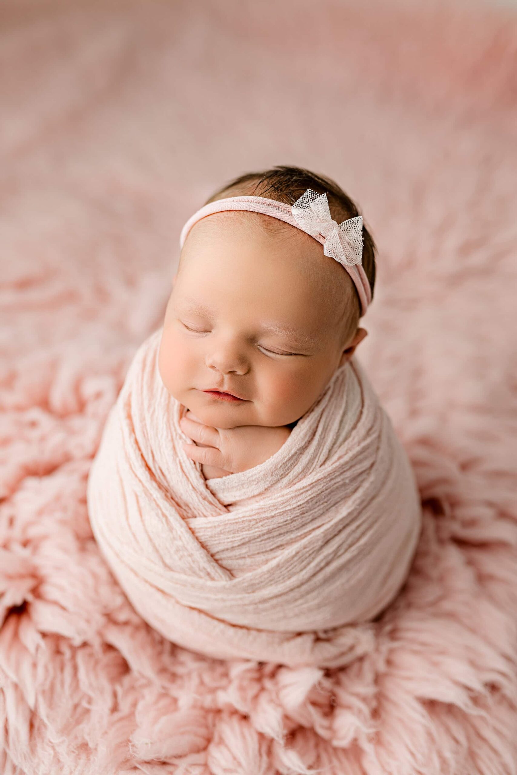baby girl is wrapped in a pink wrap with criss cross details and wearing a matching pink bow headband as she smiles on a pink fur background