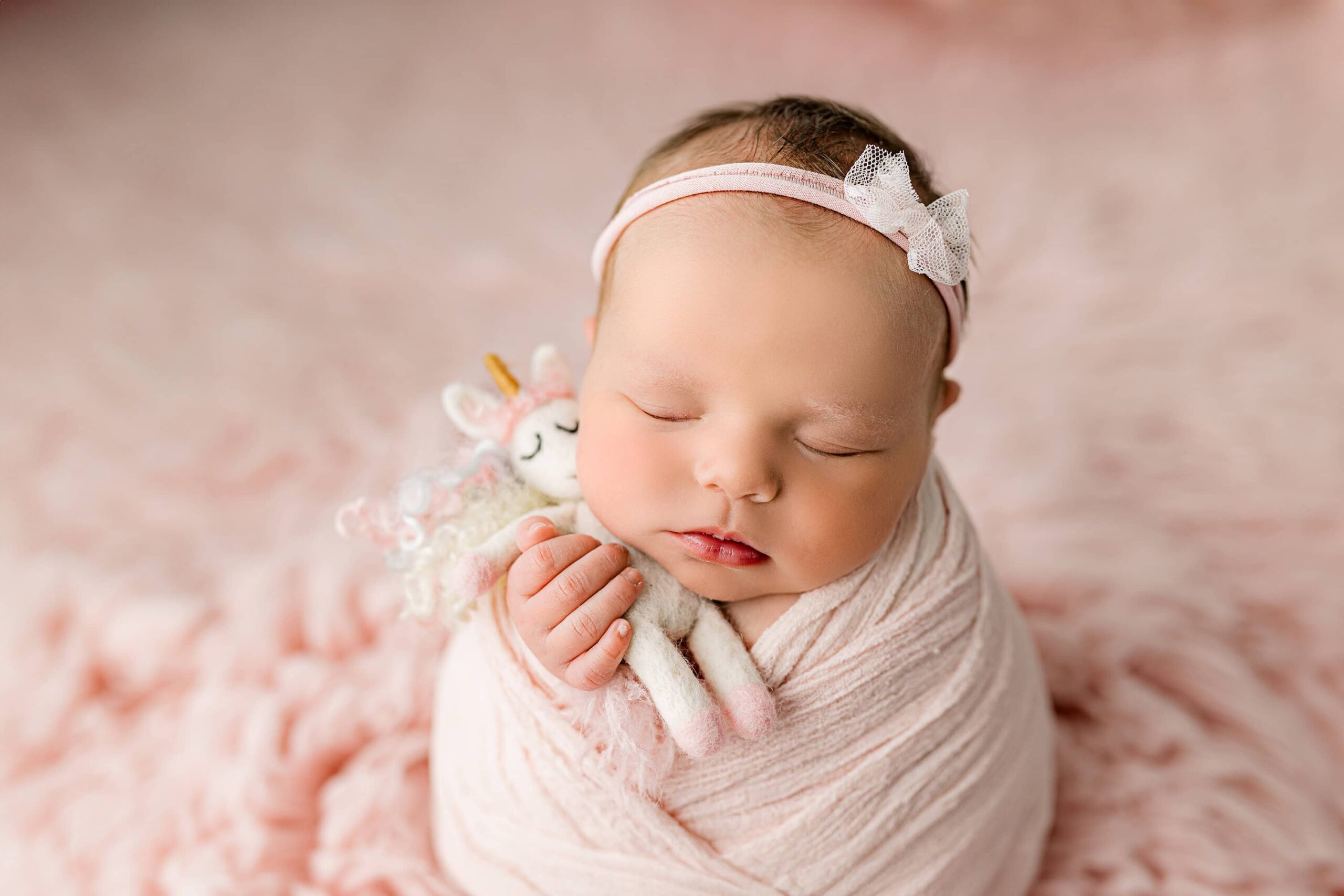 newborn baby girl snuggles with a unicorn while posed on a pink fur and wrapped in a criss cross pink wrap wearing a pink bow headband
