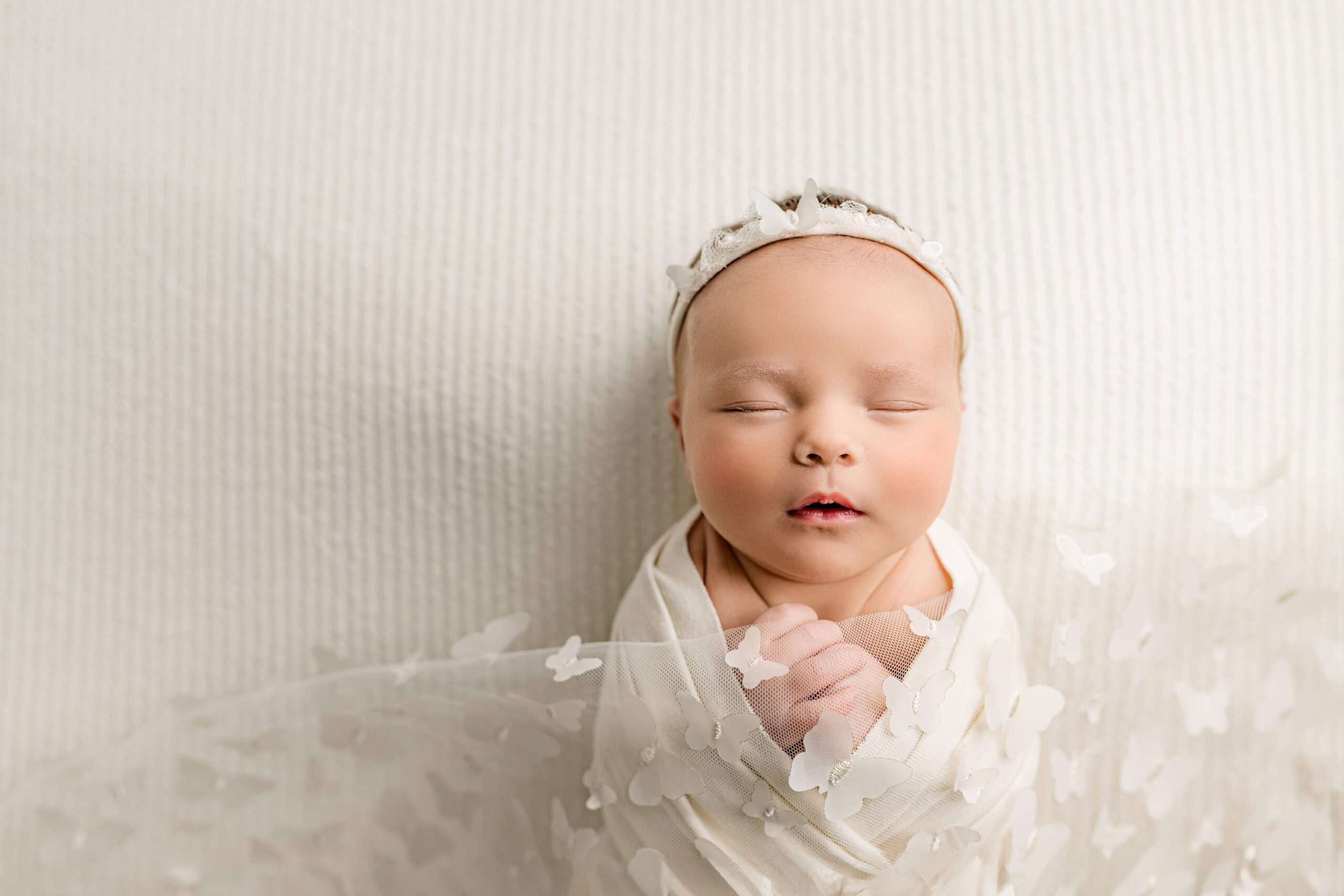 an overhead image of a newborn girl posed and wrapped in cream wearing a cream headband with butterfly trim and a butterfly overlay on her