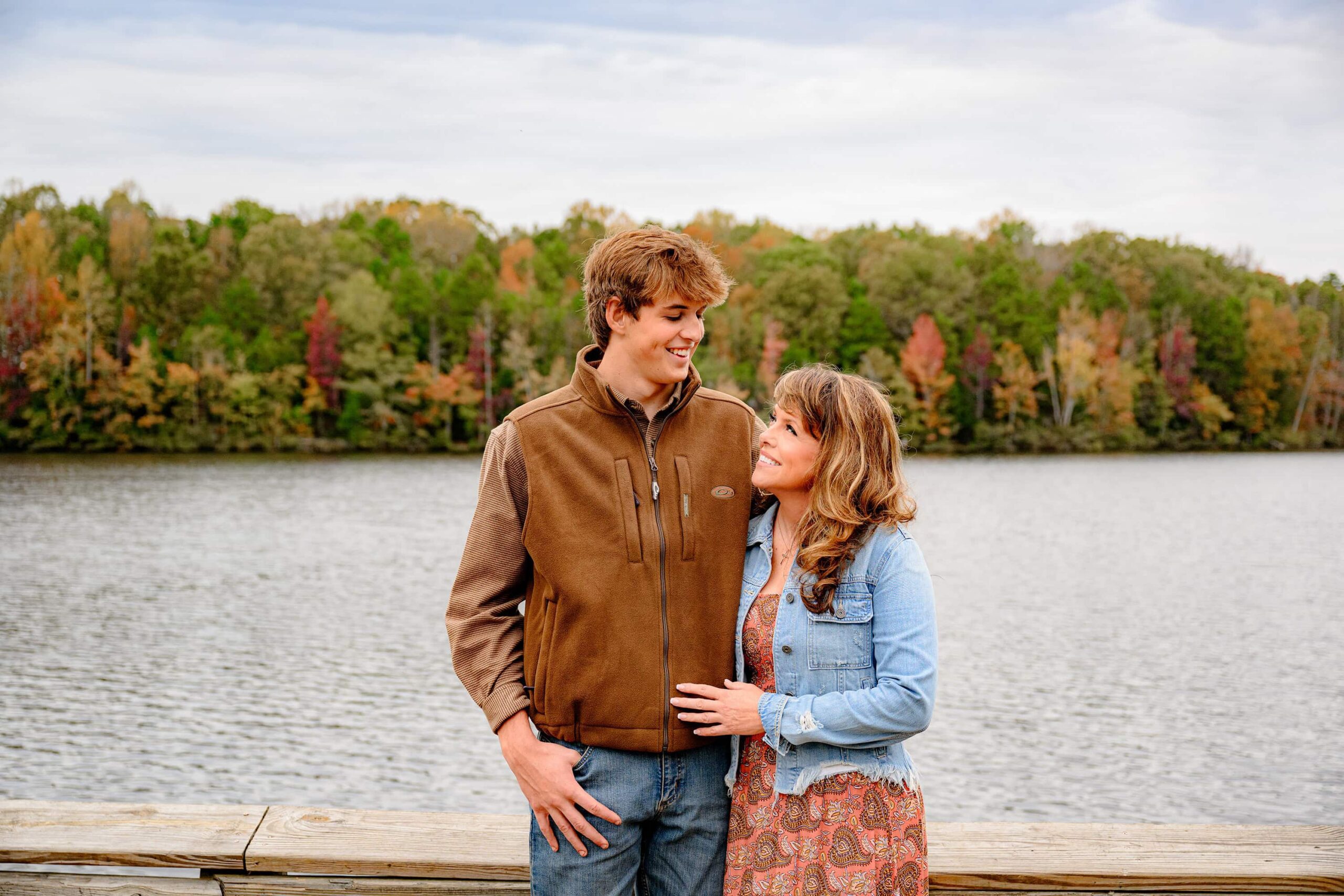 photo of a mom and her son looking at each other smiling during his senior session with fall trees and a lake behind them