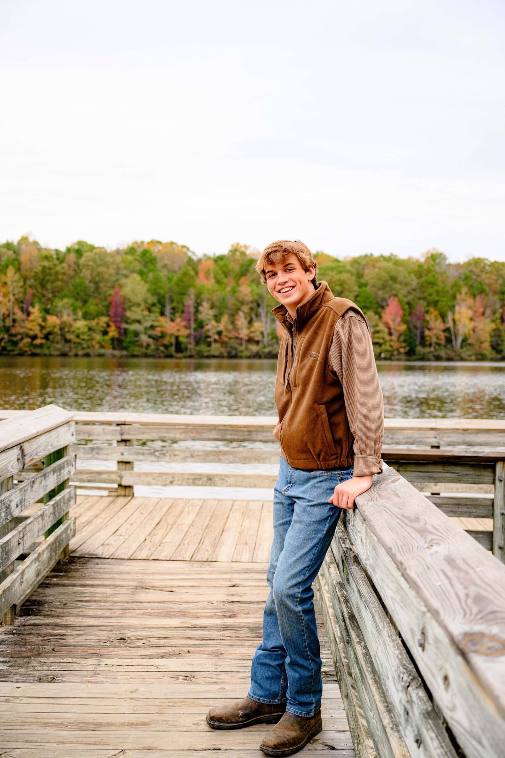 parkwood grad stands leaning one hand on a wooden railing with fall colors and a lake behind him. he is wearing a brown vest, jeans, and a brown shirt with boots