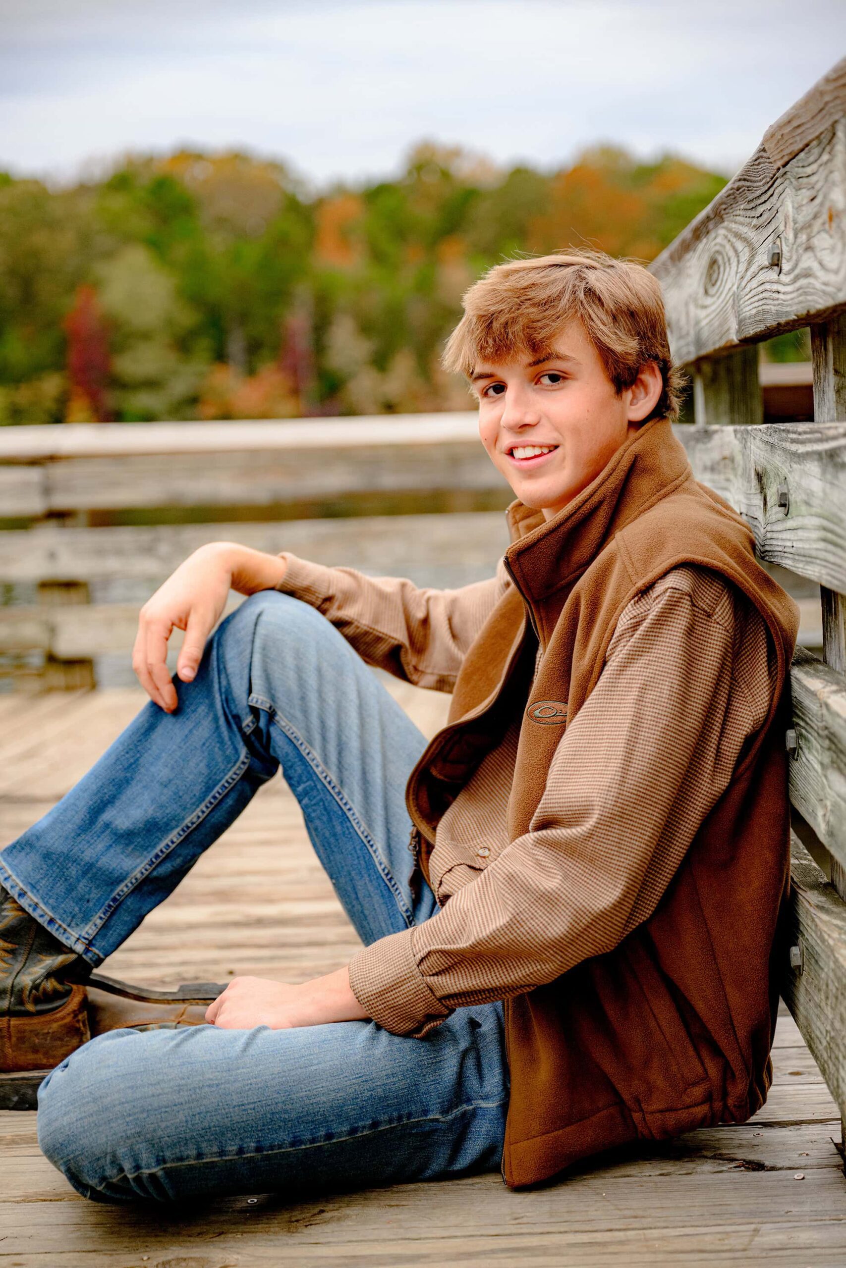 sitting against a wooden railing this charlotte area senior wears a brown vest, brown shirt, and jeans with boots