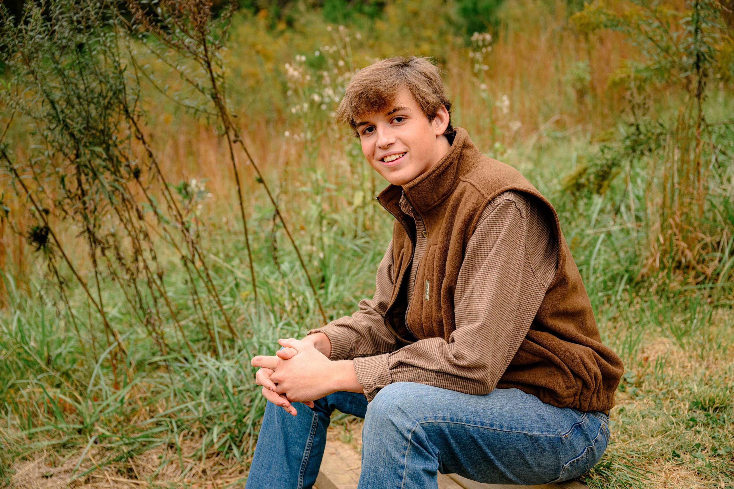 parkwood country boy sits for his senior photos in a park in lancaster, sc. he is wearing jeans, brown vest, brown shirt, and boots