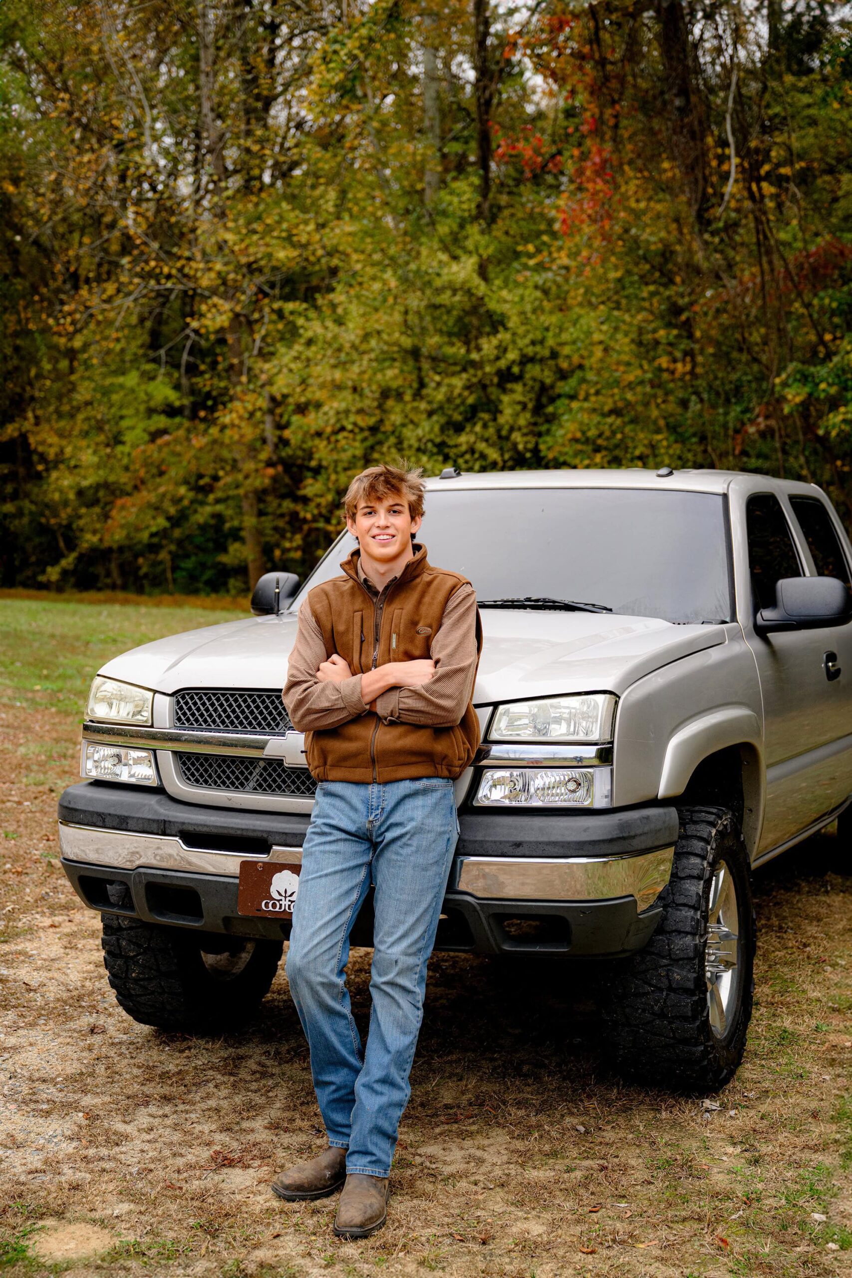 parkwood senior boy leans against his truck with his arms crossed while wearing a brown vest, brown shirt, and jeans