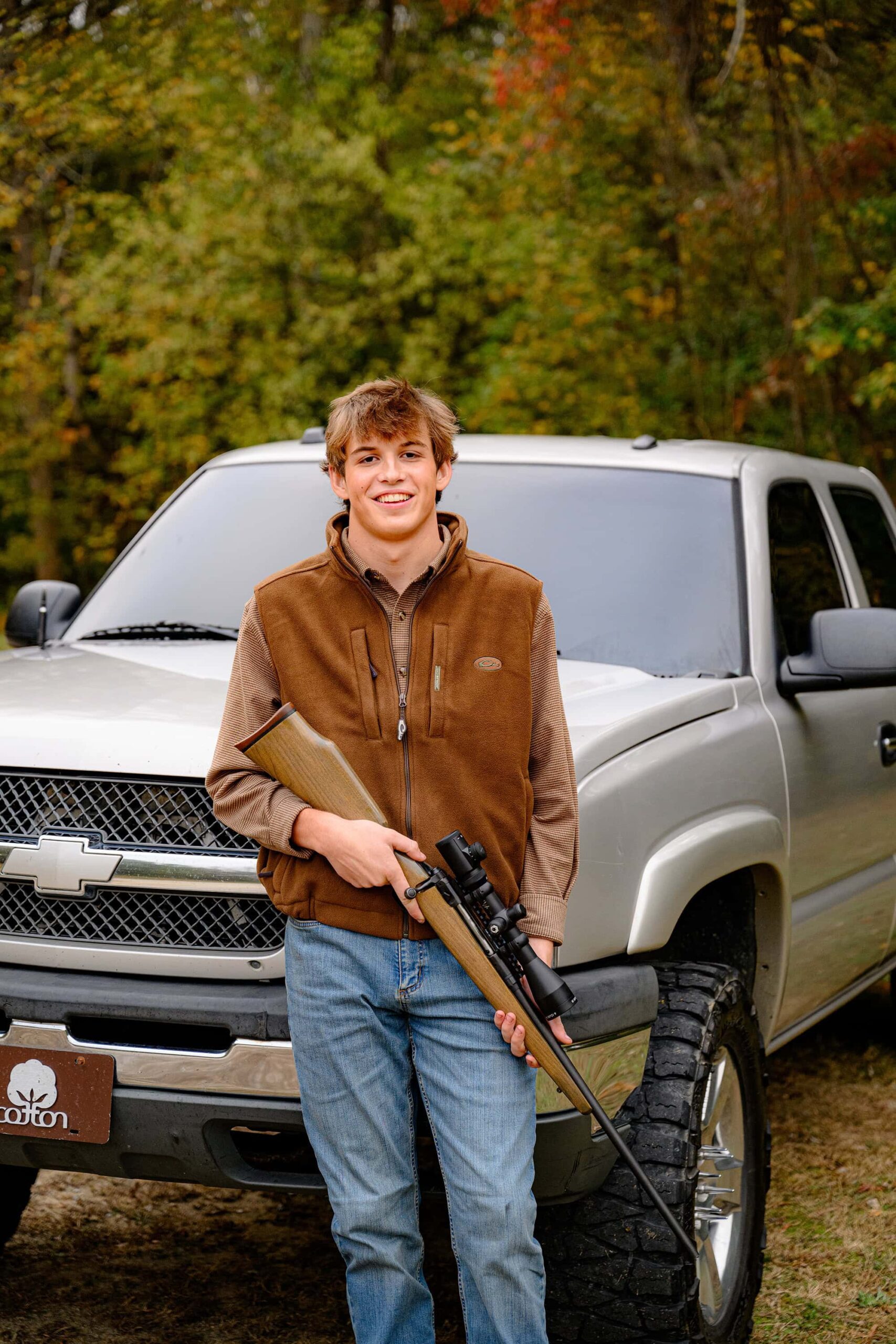 senior boy leans against his grey truck holding his hunting rifle while wearing jeans and a brown shirt and vest