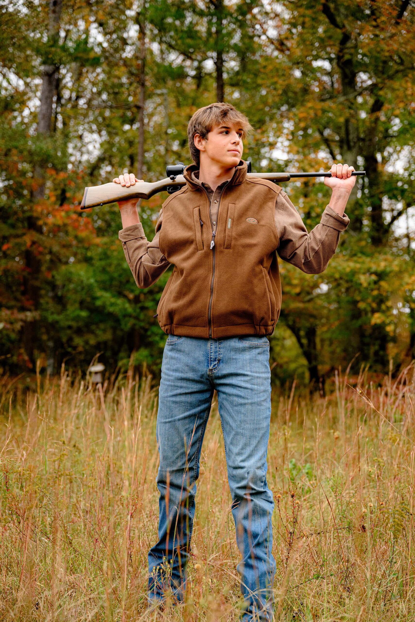 senior boy stands in a lancaster, sc field wearing jeans and a brown vest and shirt while holding his hunting rifle behind his head
