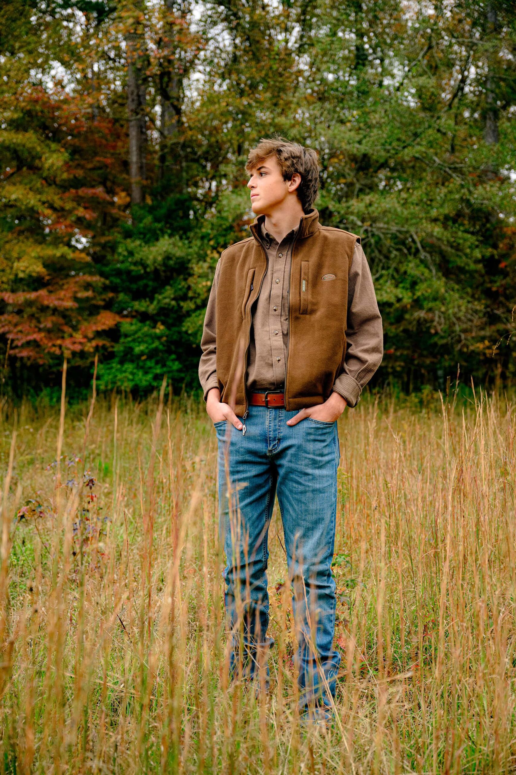 parkwood graduate stands in tall grasses wearing jeans and a brown top and vest