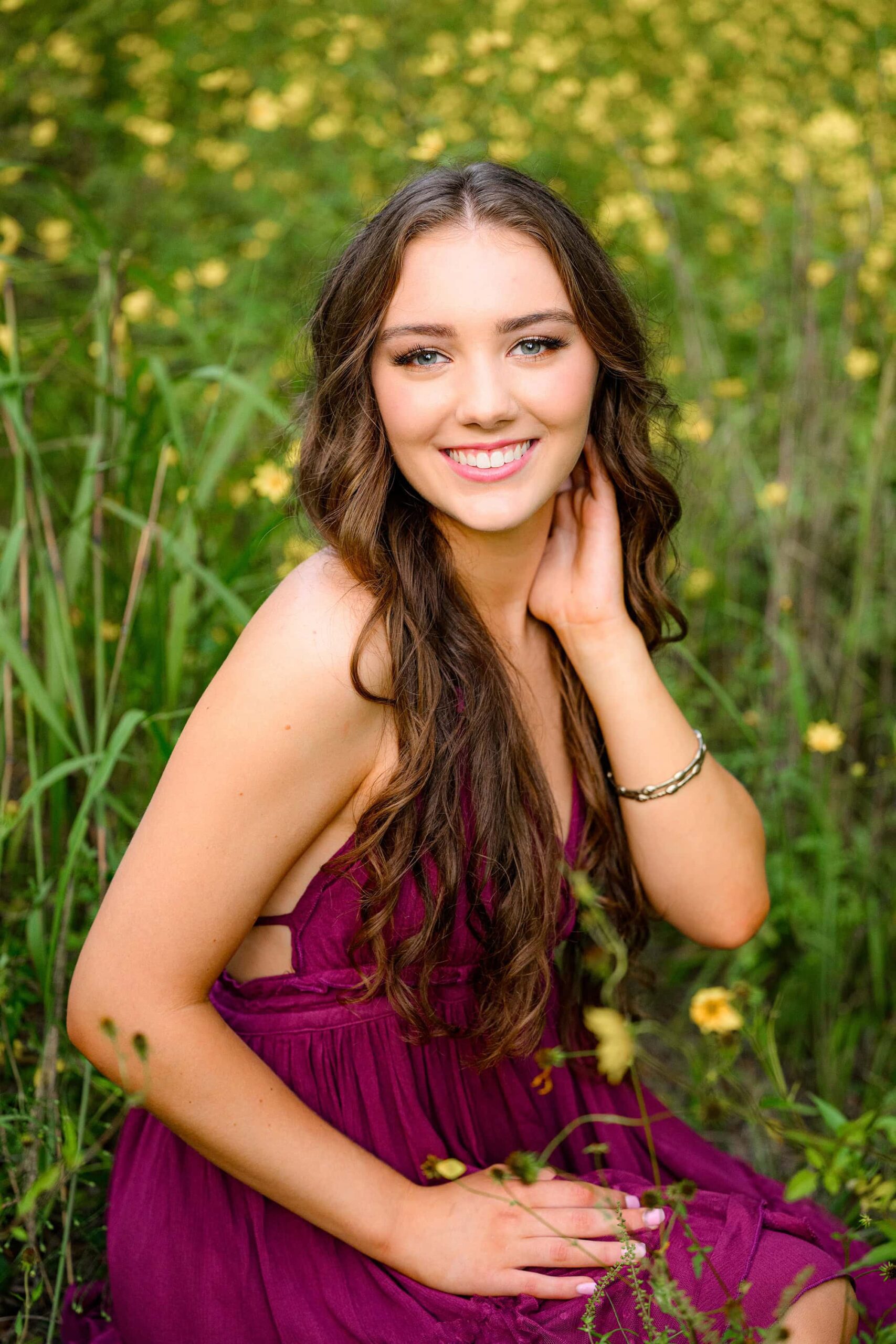 senior girl wears purple dress and sits in field of yellow blooms