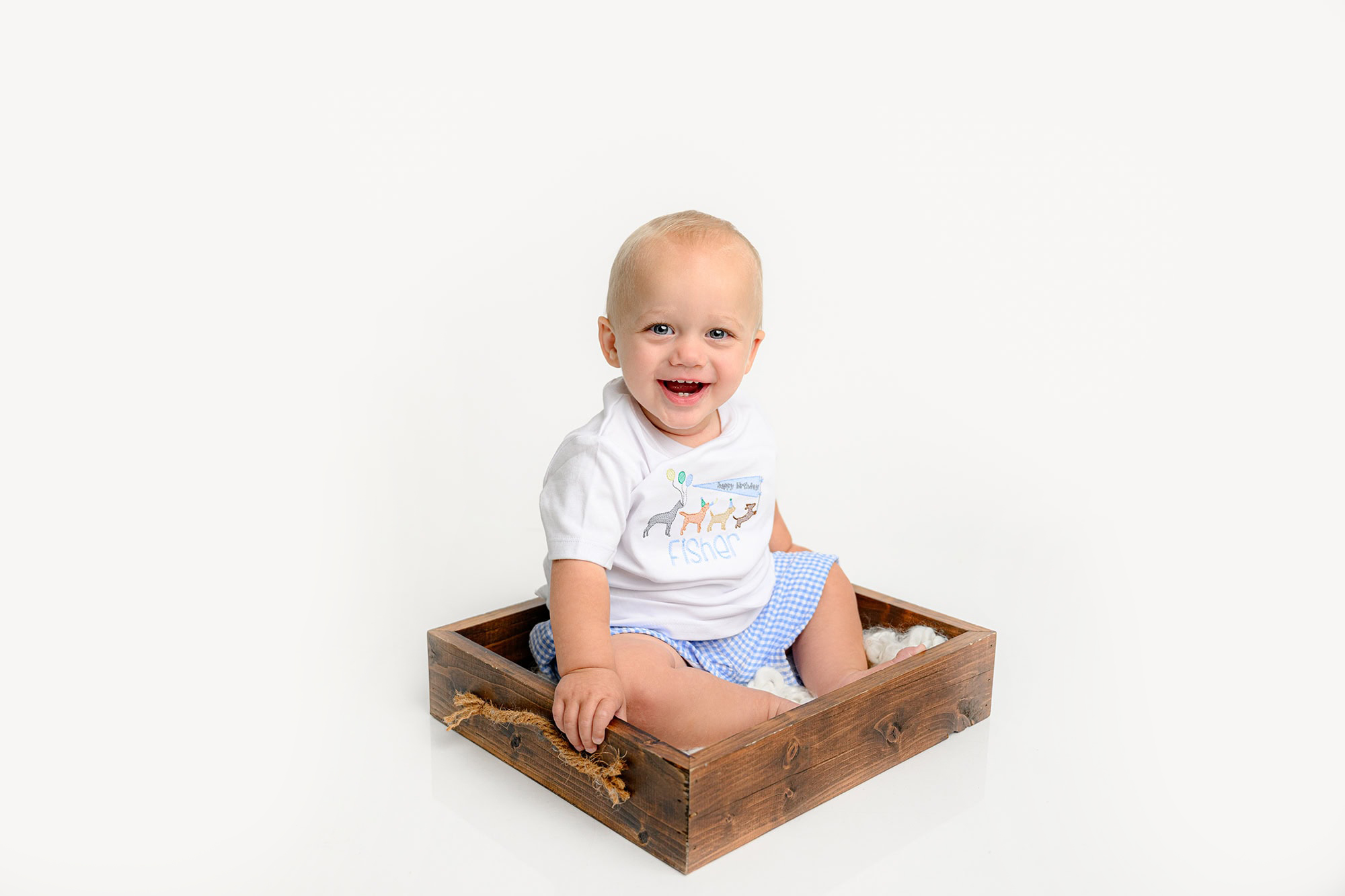 A one-year-old baby boy sits on a little brown prop wearing a birthday shirt in a simple white studio set up.