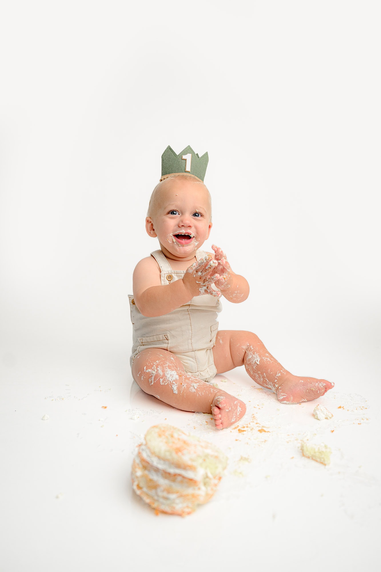 little one year old boy claps as he is covered in cake during his simple cake smash session and wearing tan overalls and a birthday hat