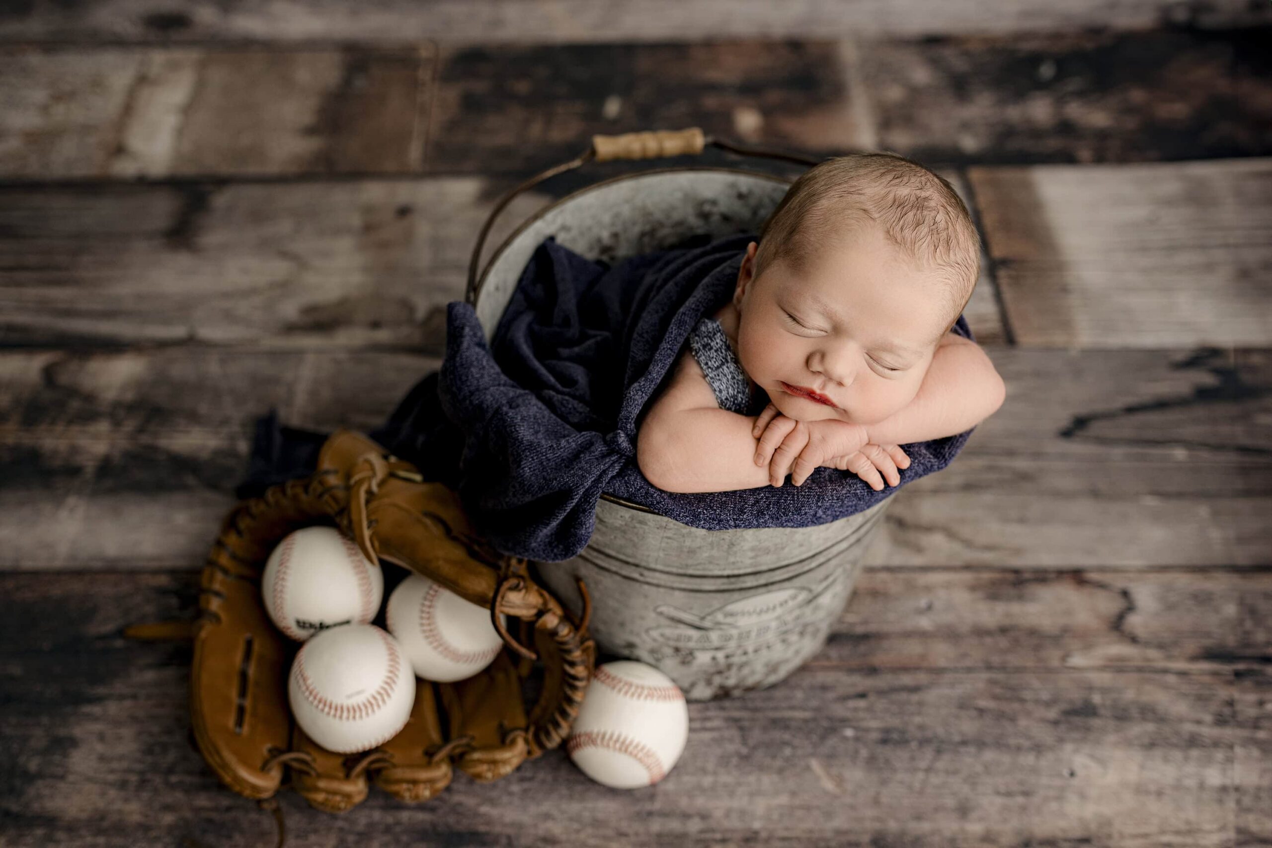 newborn baby boy posed in a bucket with baseball glove and balls beside the bucket