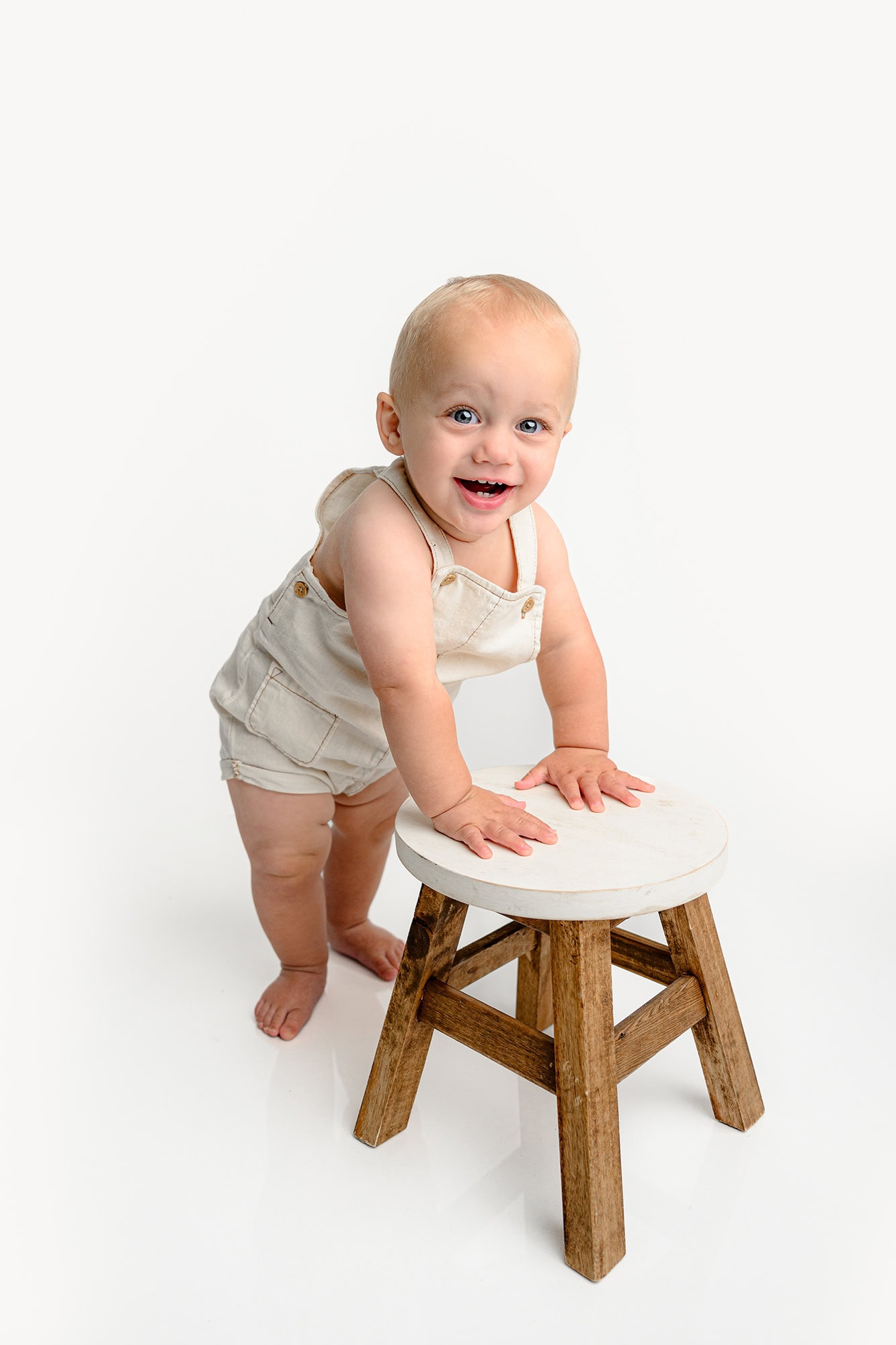 little boy stands holding onto a stool wearing cream overalls