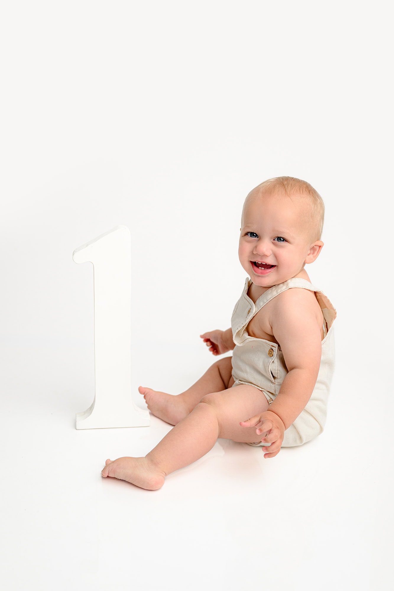 little boy sits on a simple white studio background during his birthday photoshoot beside a large number one prop wearing tan overalls