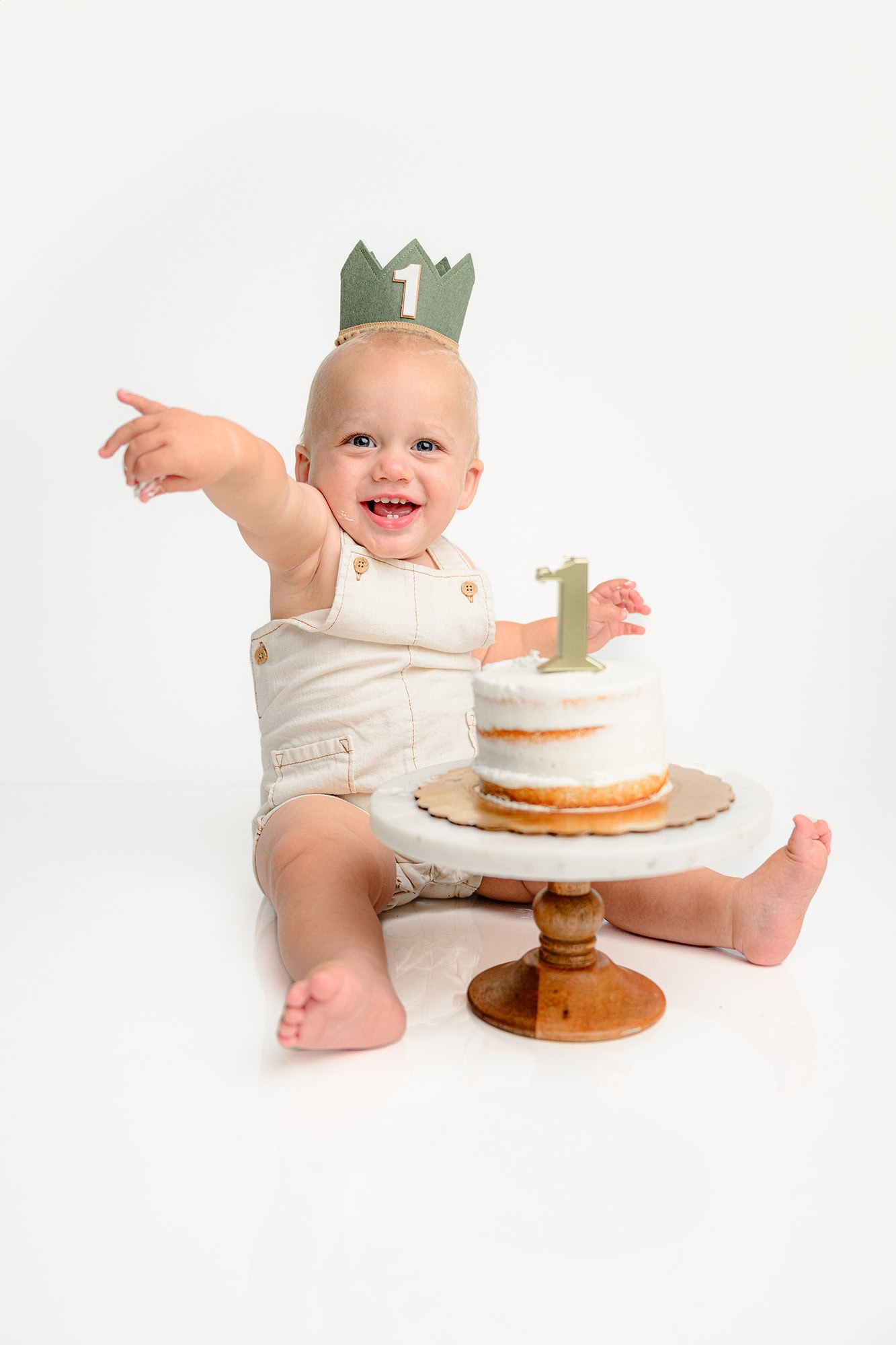 little boy sits in cream overalls sitting behind his cake on a stand during his birthday photoshoot