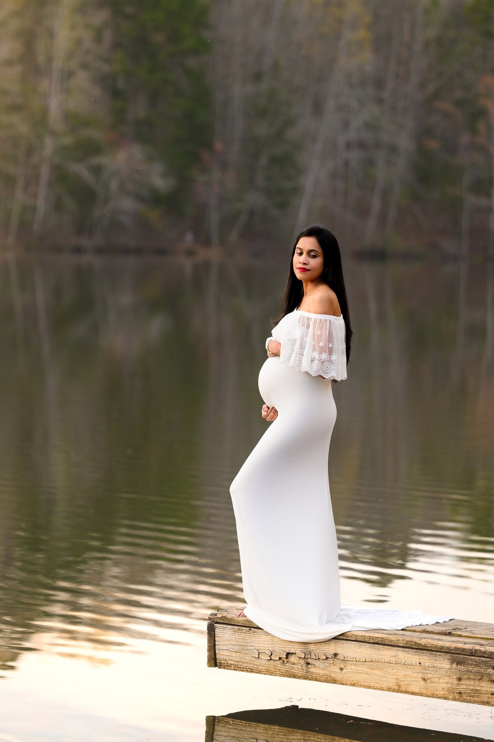 expectant mother with long dark hair and red lipstick poses wearing a white maternity dress with a boho collar beside a lake in south carolina