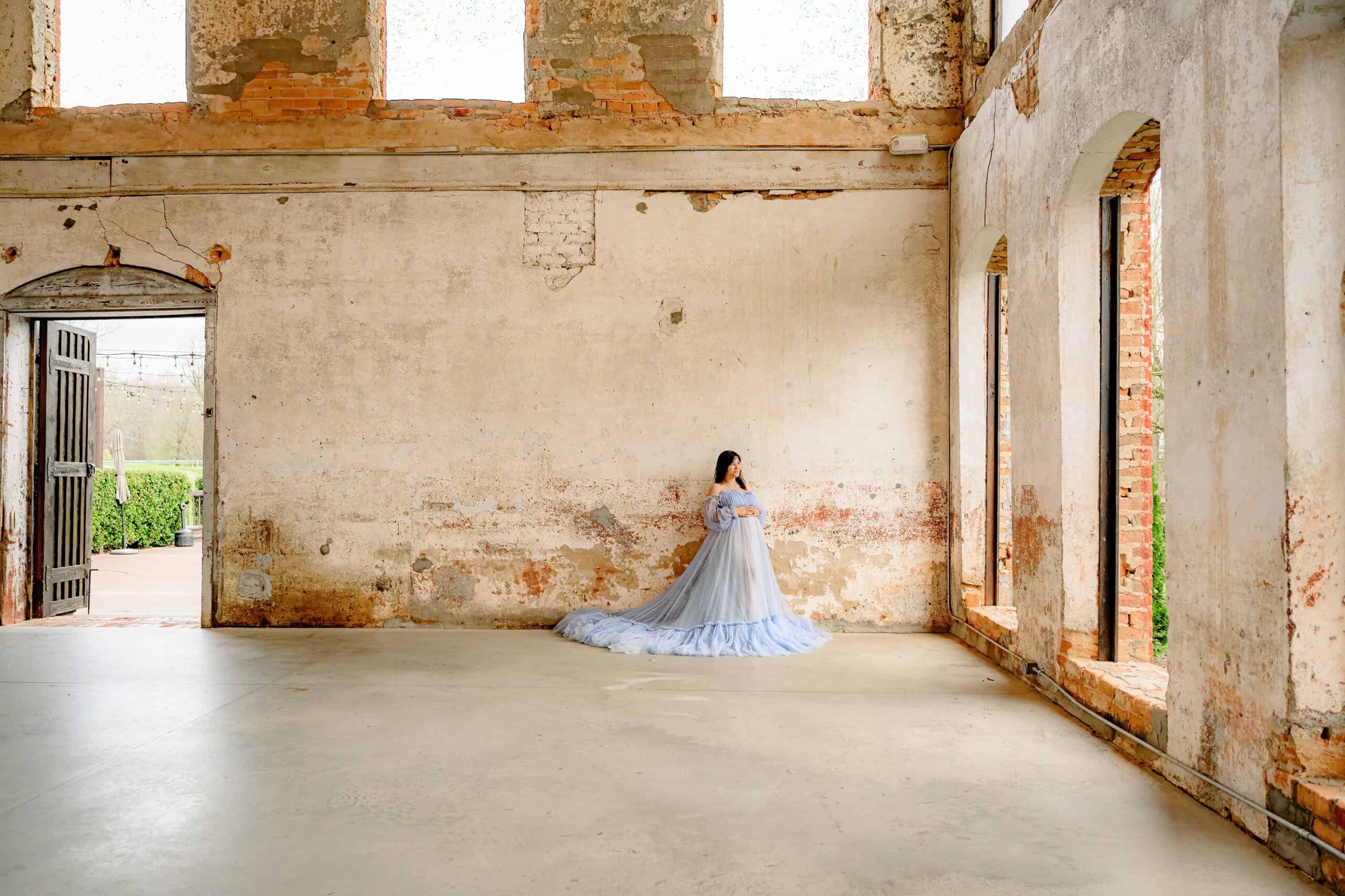 expectant mother dressed in a blue dress stands beside a historic wall at the providence cotton mill