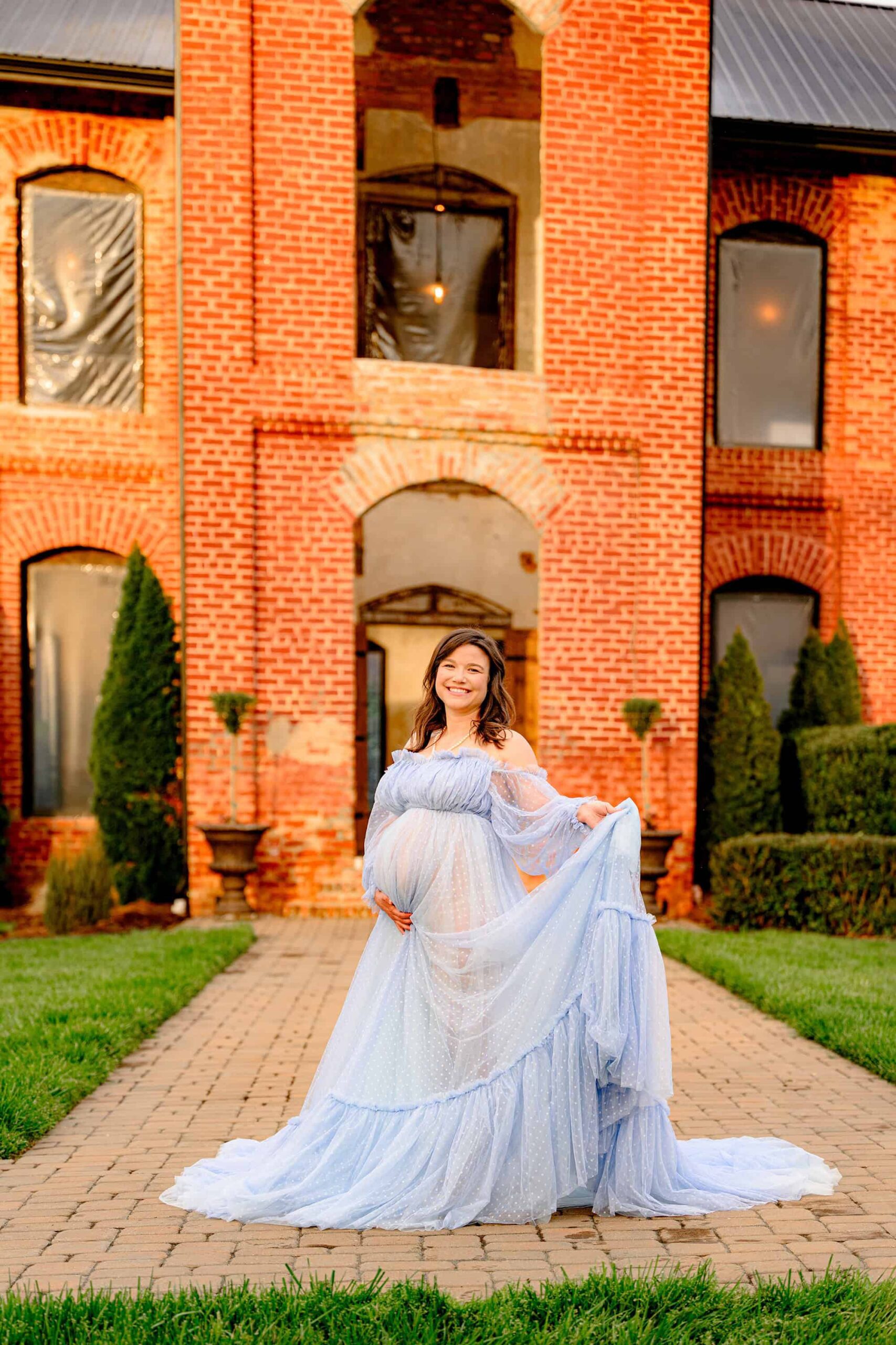 pregnant mother poses holding the train of her blue maternity dress with the historic providence cotton mill behind her