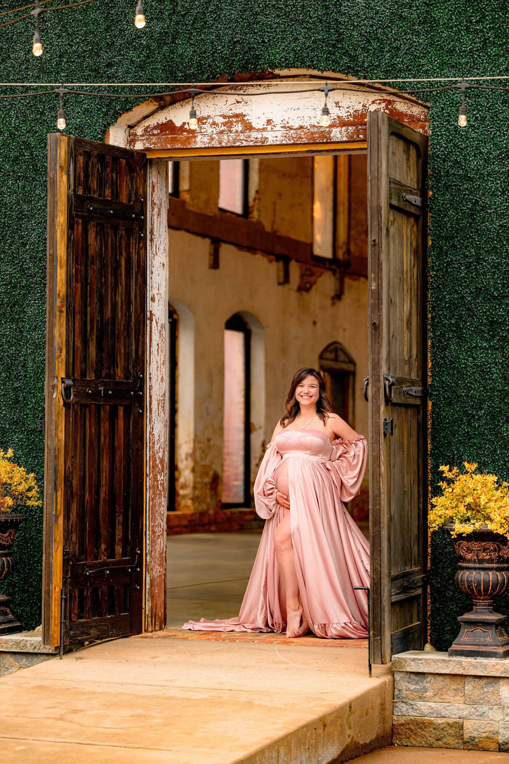 pregnant mother stands behind two large doors at this exclusive venue wearing a pink satin dress and holding her belly