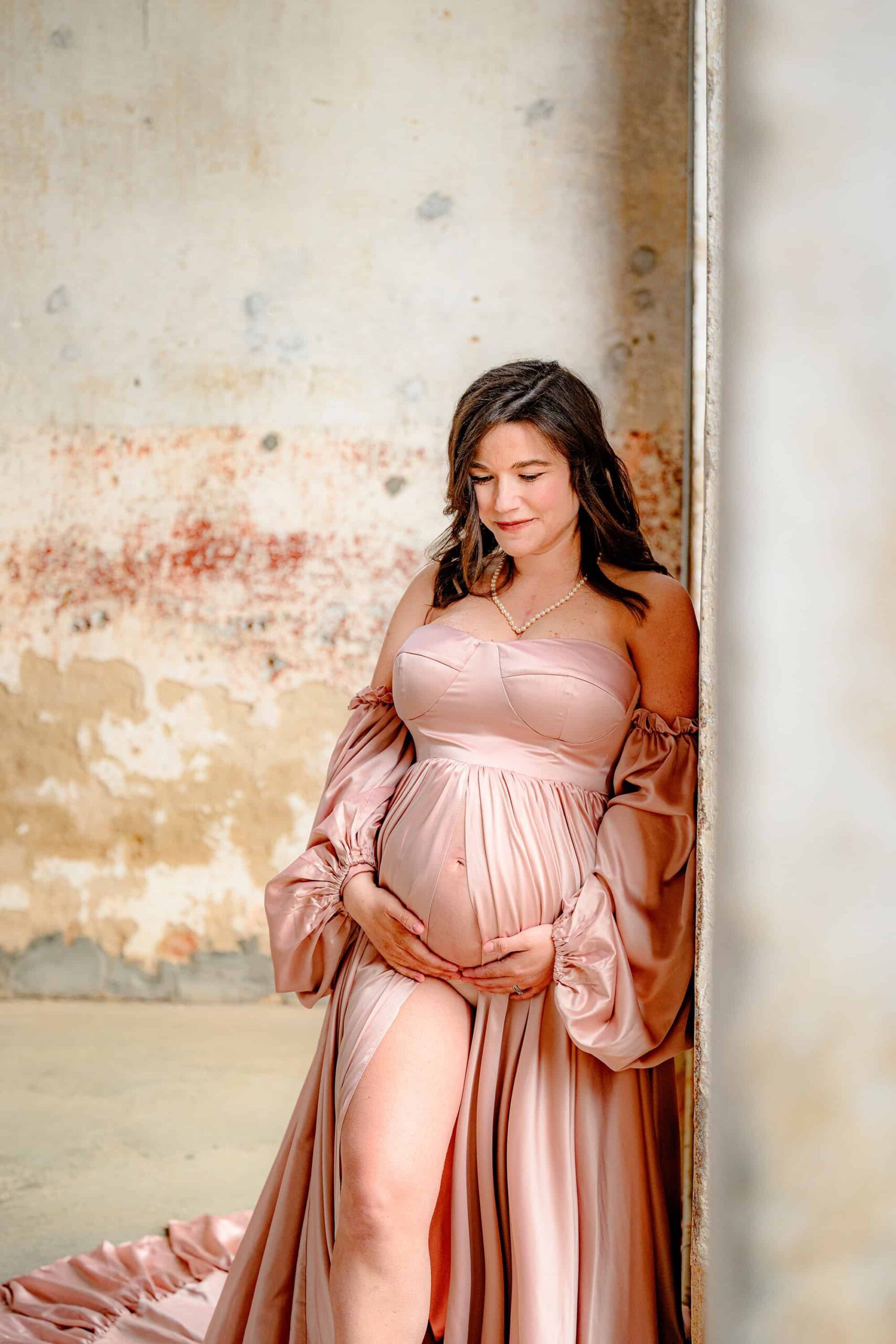 expectant mother looks down at her belly during her maternity photoshoot while leaning against a wall