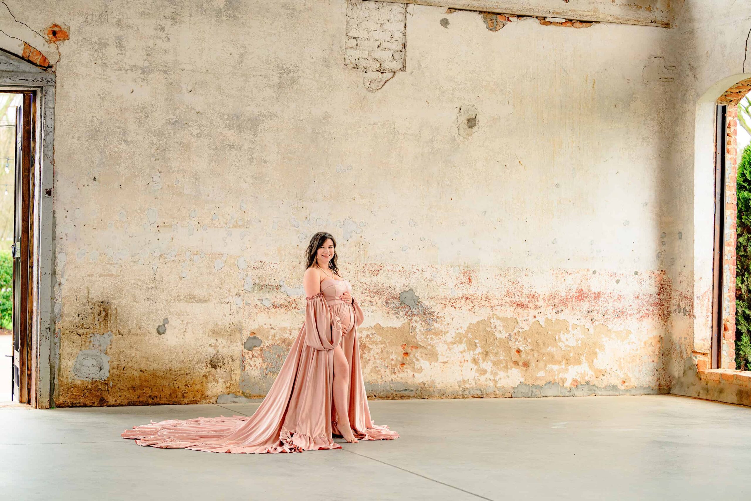 expectant charlotte mother stands in front of a historic wall at the providence cotton mill during her maternity photoshoot wearing a satin pink dress