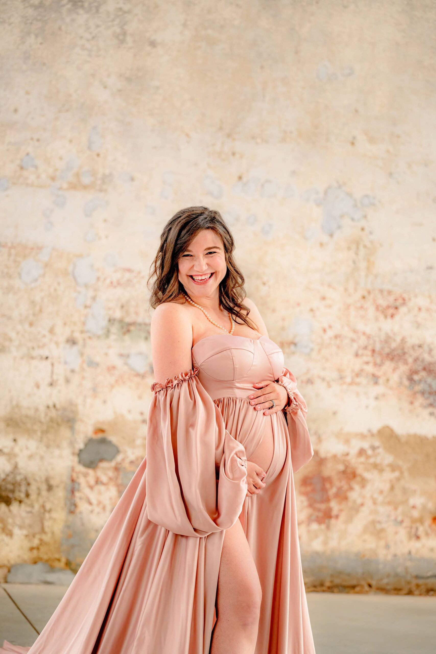 charlotte mother laughs as she poses touching her belly during her pregnancy photoshoot wearing a pink satin dress
