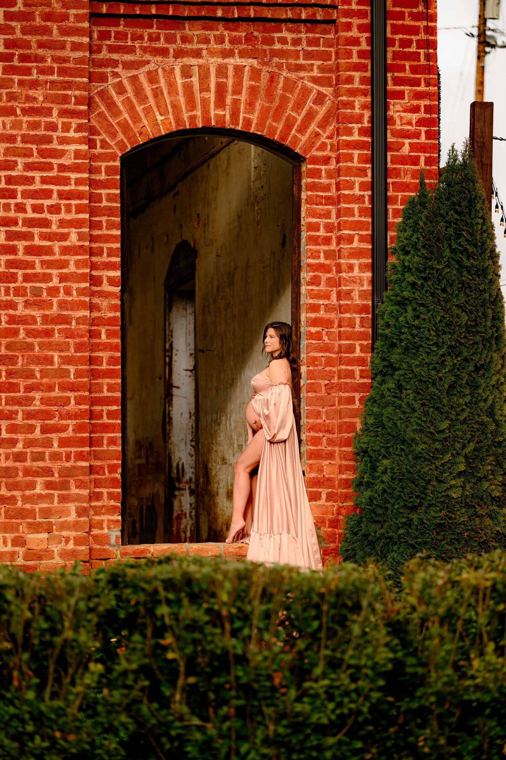 Charlotte expectant mom dressed in a pink satin dress holds her belly as she is posed in a window for her maternity photoshoot at the providence cotton mill