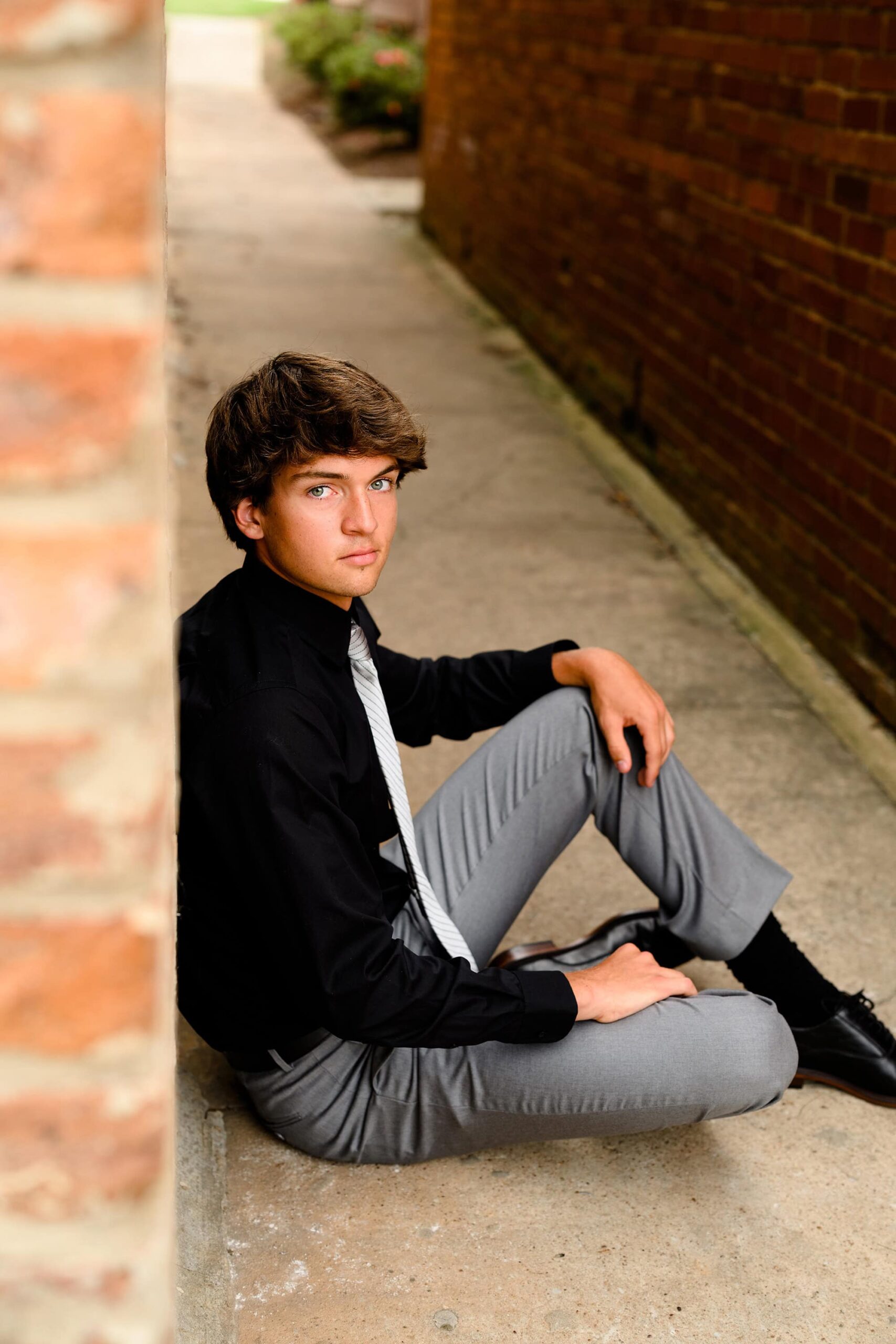 Union County Senior sits in grey dress pants and black shirt with grey tie for senior portraits in downtown waxhaw