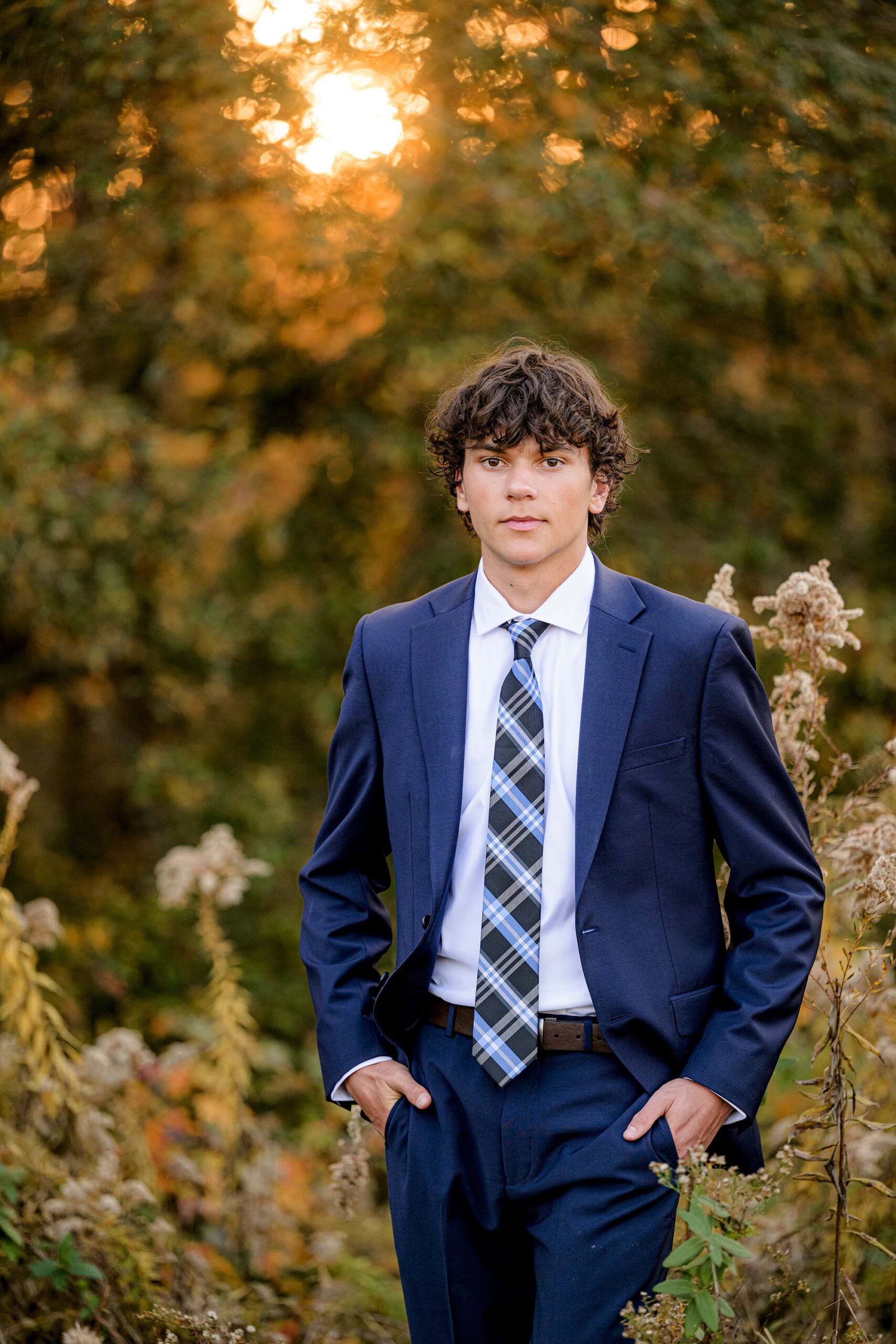 senior portrait of a boy in a navy suit with a navy plaid tie standing in a field with golden light behind him