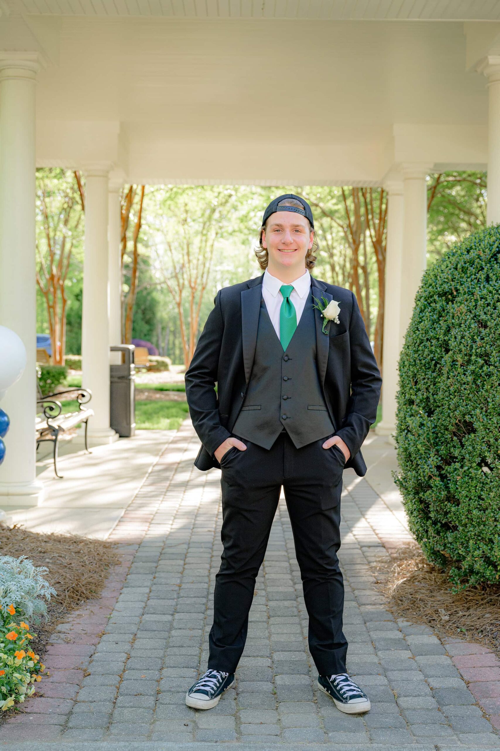 senior boy stands under pavilion for prom photo in black suite with green tie and hat