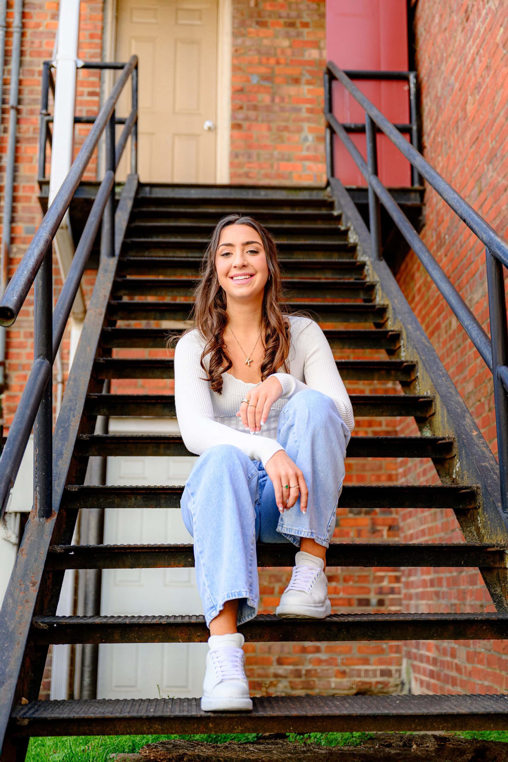 senior sits on a metal stairway wearing jeans and a white top with white sneakers in downtown waxhaw for her grad photoshoot