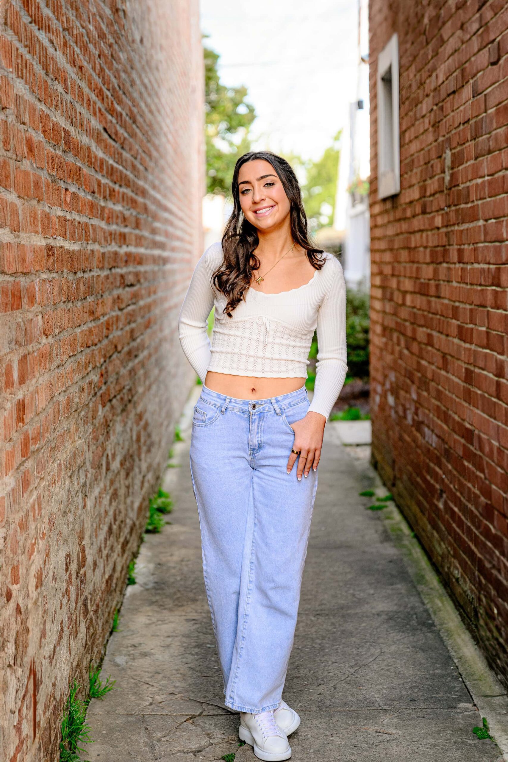 senior poses with her hands in her pockets wearing jeans and a white top in a brick alleyway in downtown waxhaw