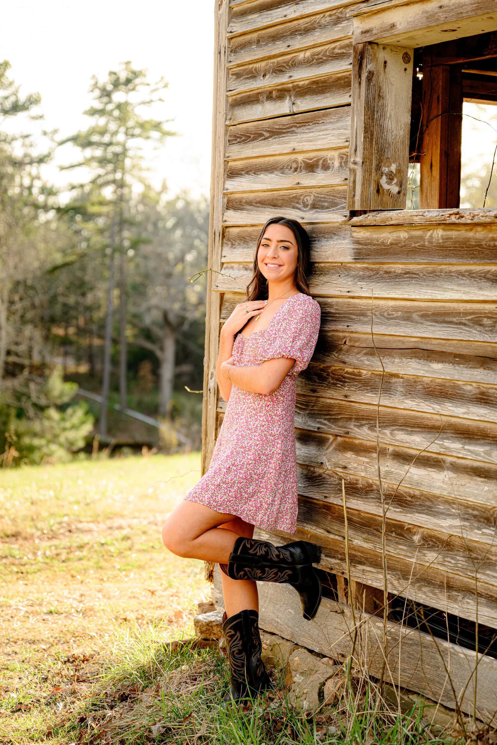 senior leans against a barn with one foot up on the barn wall. she is wearing brown cowboy boots and a floral baby doll dress