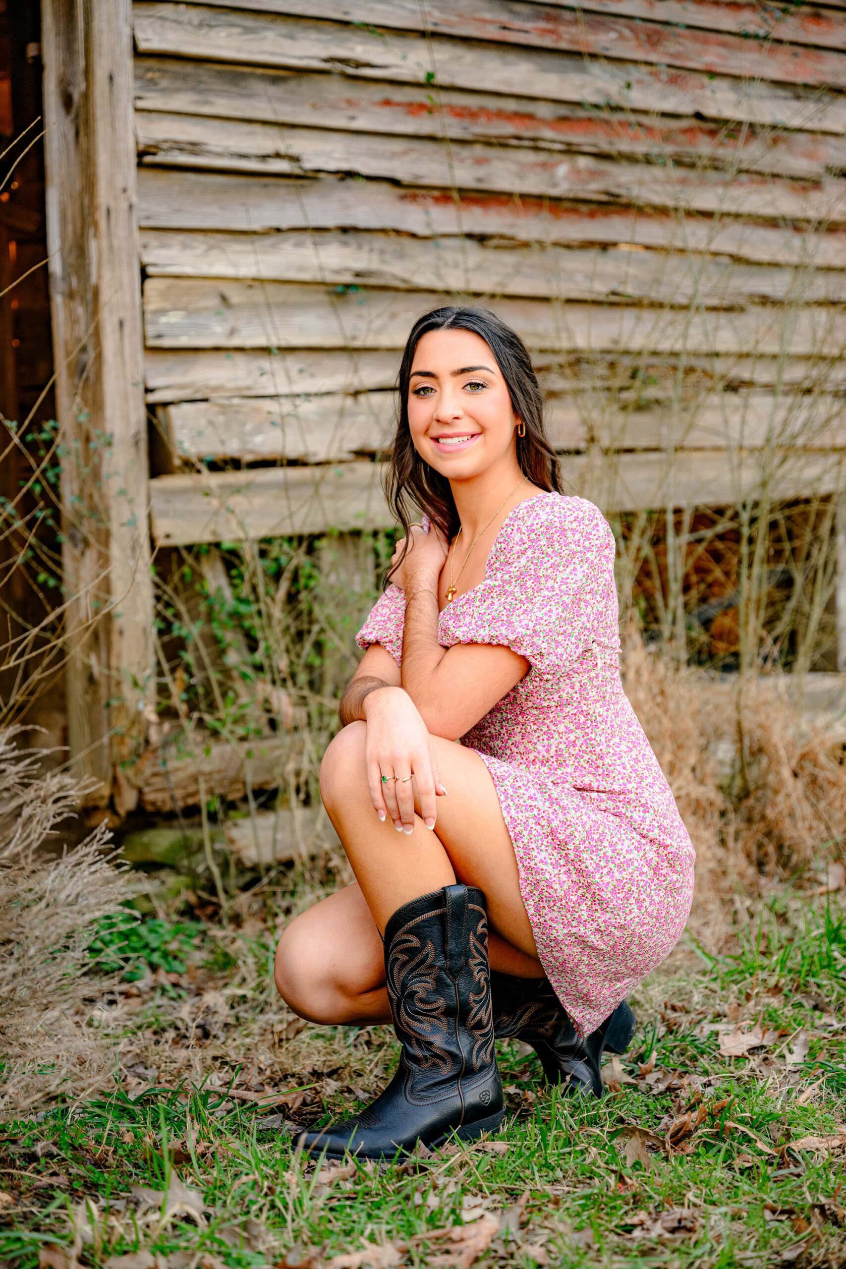 marvin ridge grad phototshoot with a senior squatting down beside a barn wearing her brown cowboy boots and floral dress