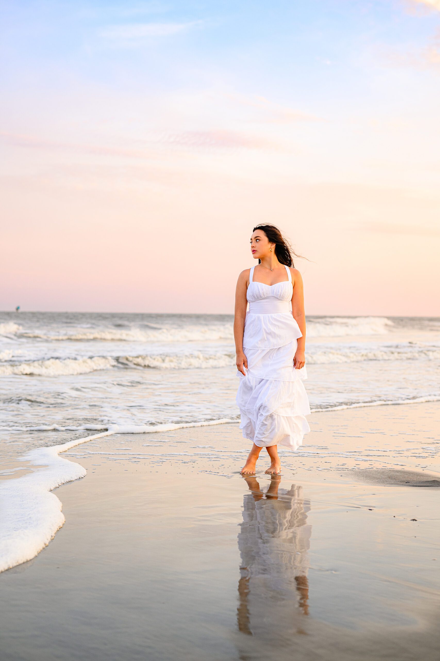 graduate stand on beach in charleston wearing a white dress looking toward the waves