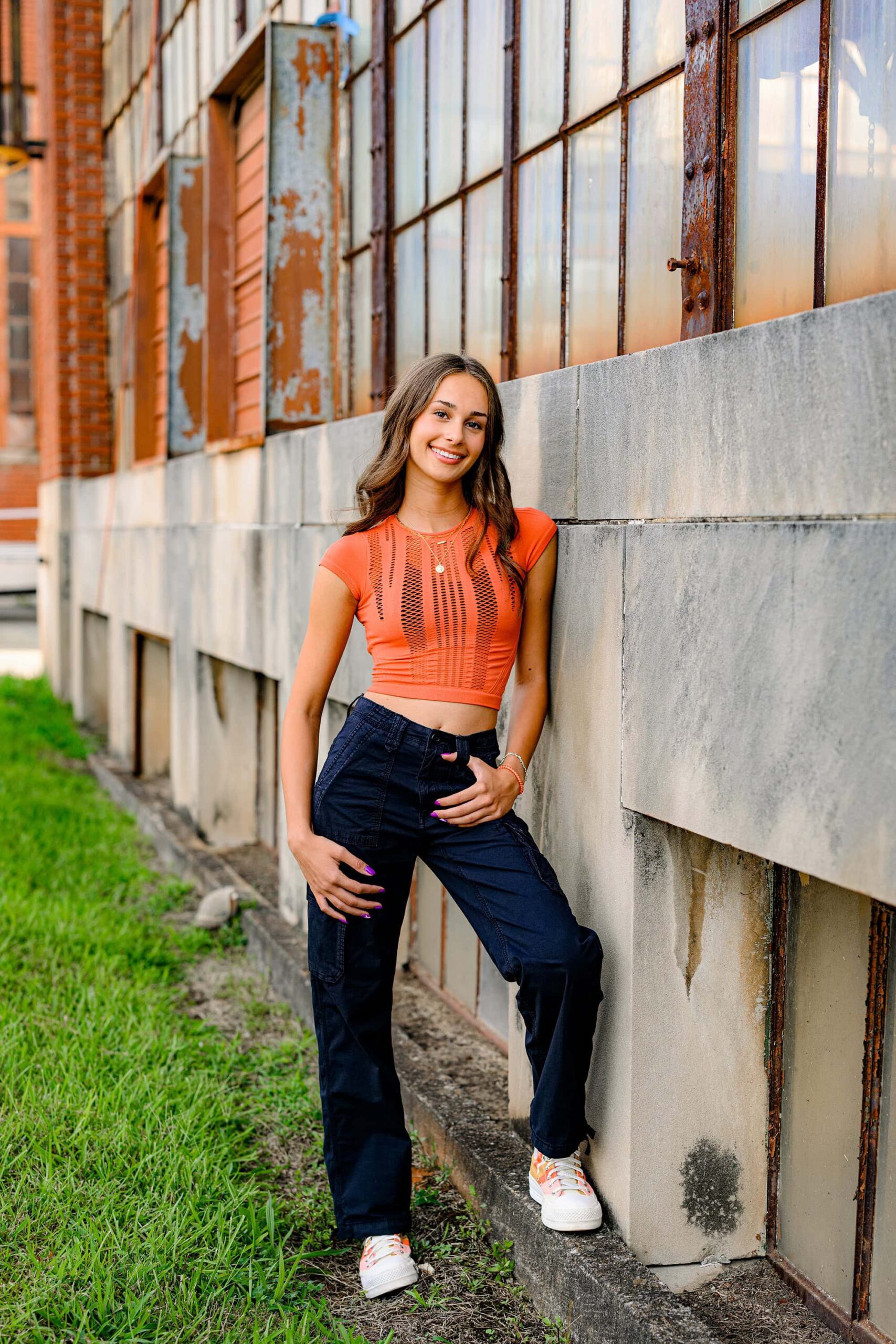 Charlotte senior poses against a wall wearing black pants and an orange top with orange and white converse shoes