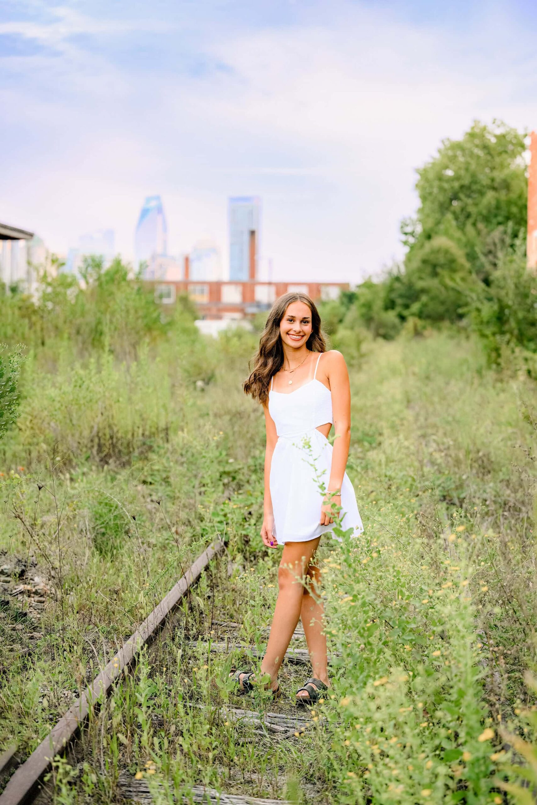 ardrey kell high school senior with dark hair stands on train tracks with charlotte skyline behind her. she is wearing a white dress
