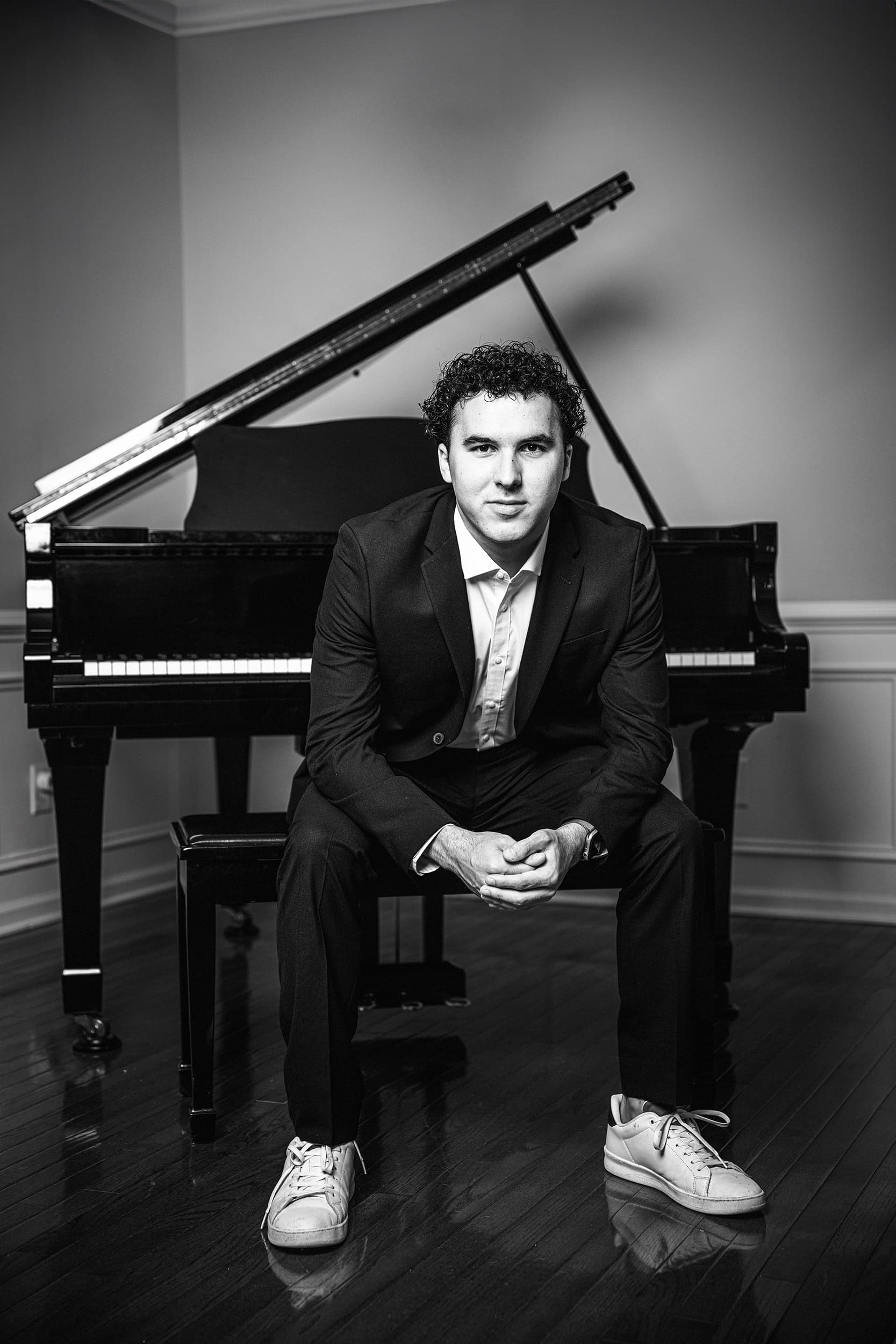 Black and White image of a senior in a suit jacket sitting on his piano bench with his piano behind him and leaning forward